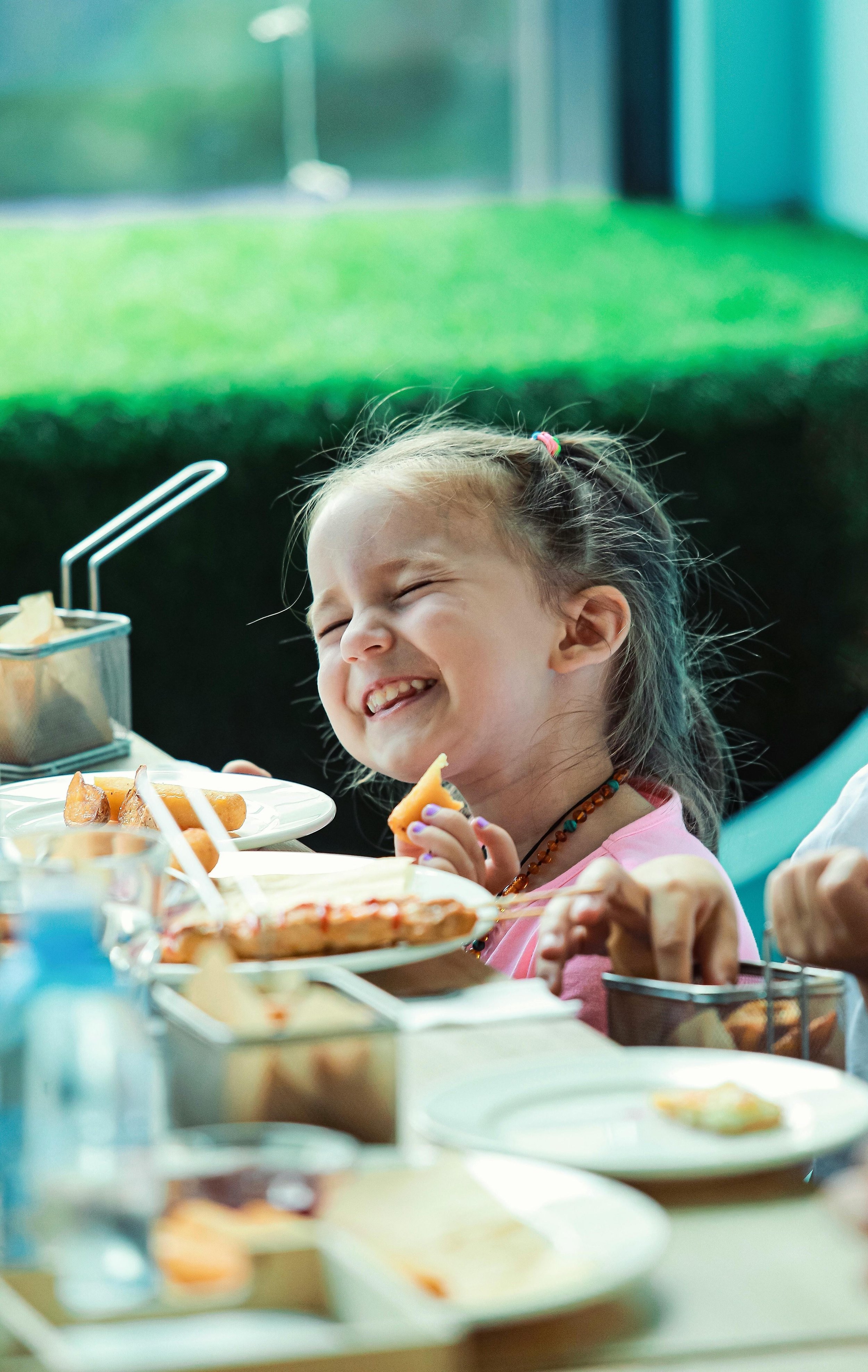 Young girl laughing at a dining table with plates of food, holding a piece of food in her hand.