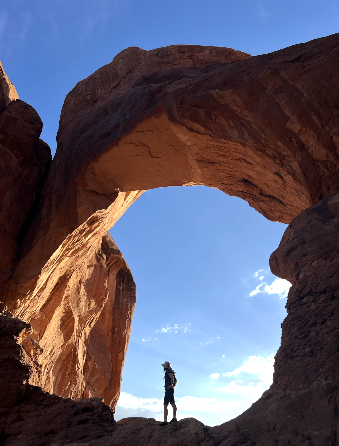 Hana wearing a hat and backpack standing at the base of a large natural sandstone arch against a blue sky with some clouds on a hike.
