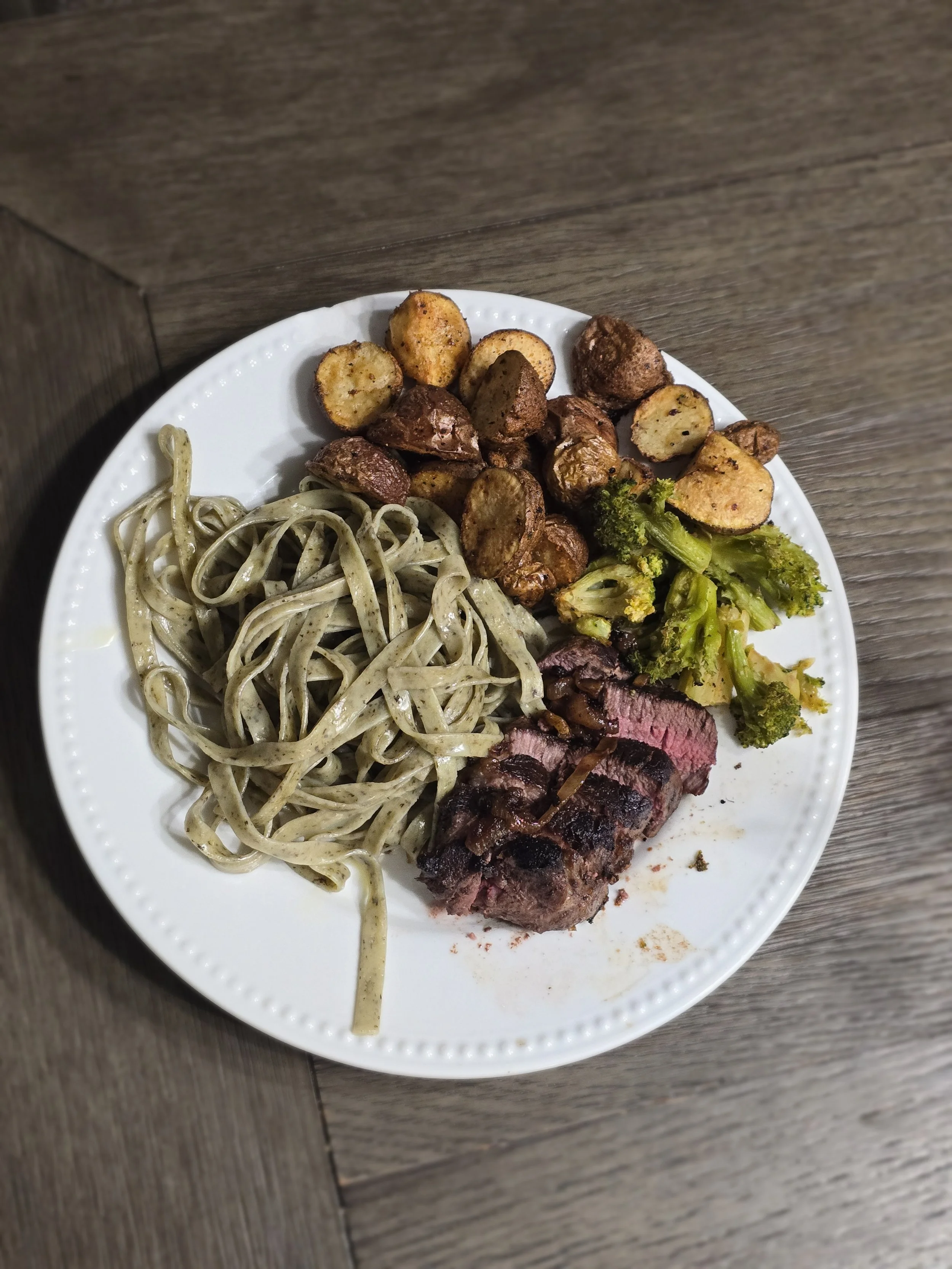 Plate with cooked steak, roasted potatoes, pasta with green sauce, and steamed broccoli.
