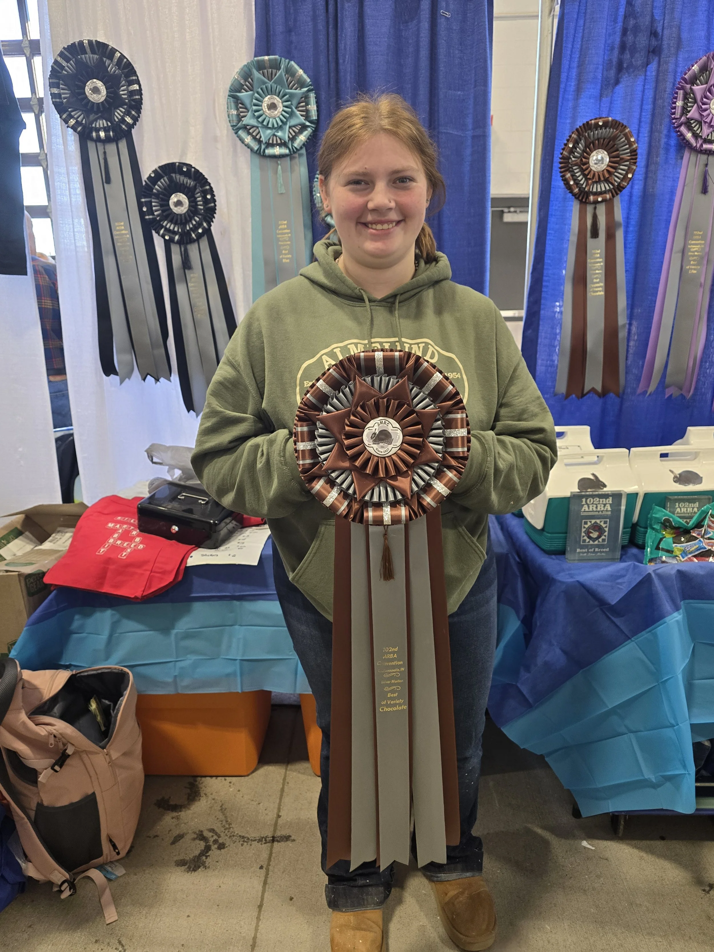 A young woman with red hair and a green hoodie holding a large award ribbon at a competition or show, with other ribbons and display tables in the background.