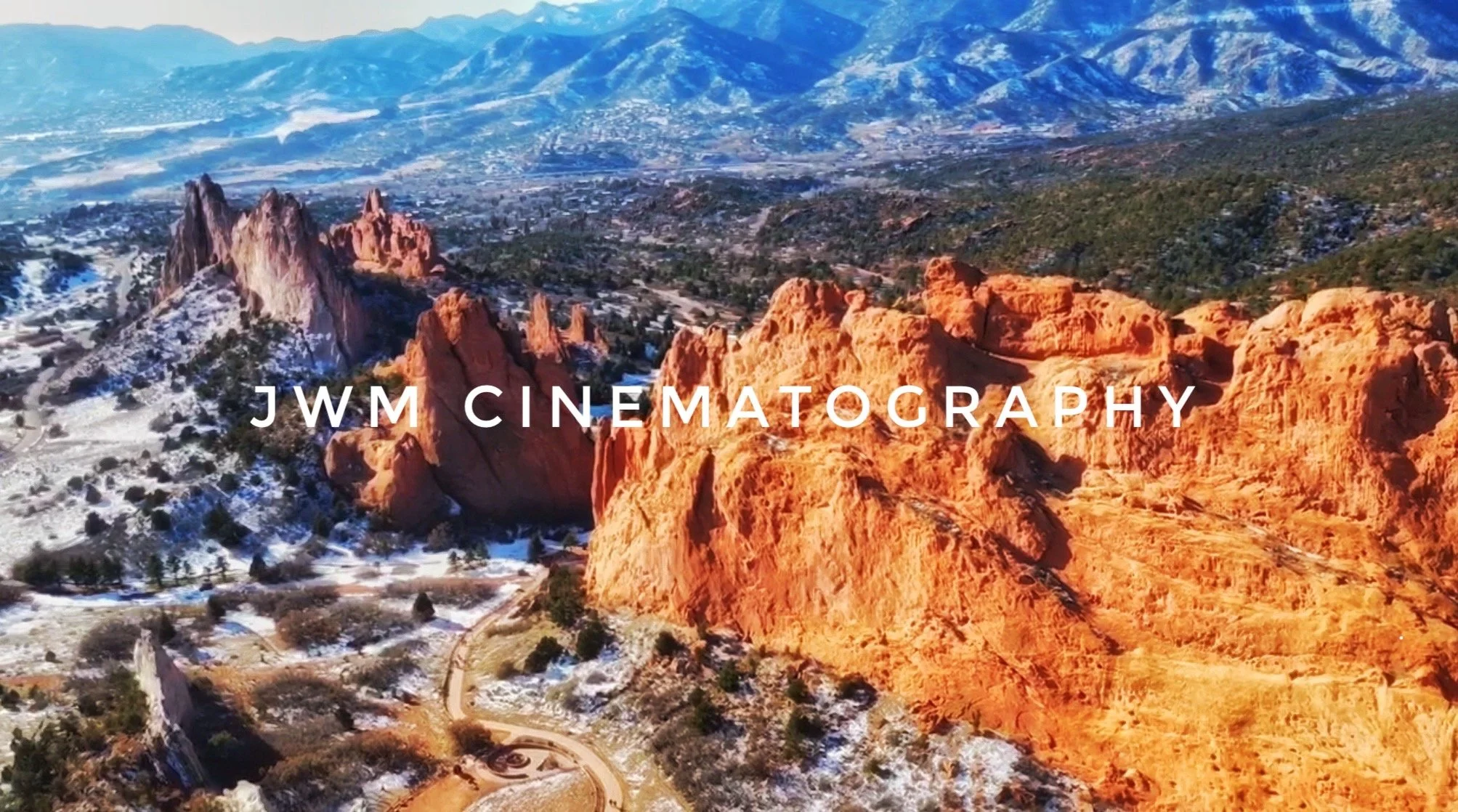 Aerial view of red rock formations in Garden of the Gods, Colorado, with snow-dusted landscape and mountains in the distance. "JWM Cinematography" text overlay.