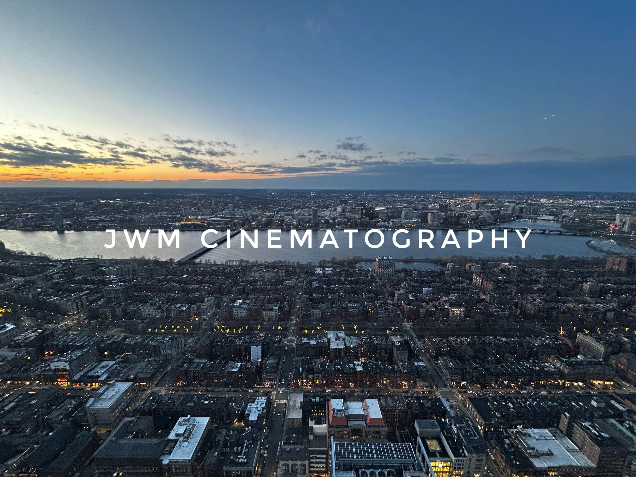 Aerial view of an urban landscape at sunset with a river running through the city. The sky is partly cloudy with an orange glow on the horizon. The text "JWM Cinematography" is superimposed on the image.