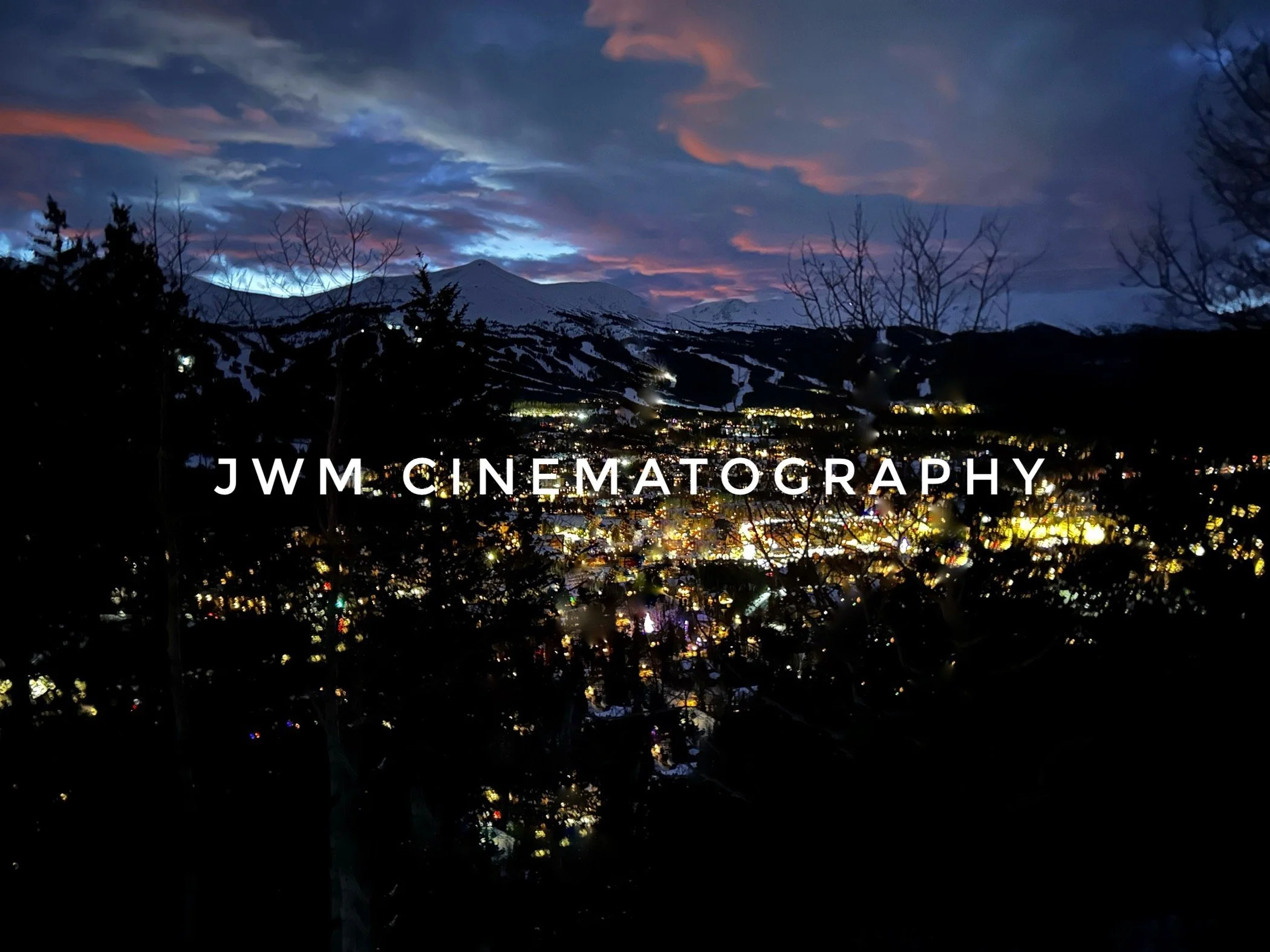 Night view of a cityscape with bright lights, silhouetted trees in the foreground, and mountains in the background under a colorful sunset sky. Text "JWM Cinematography" is visible in the center.