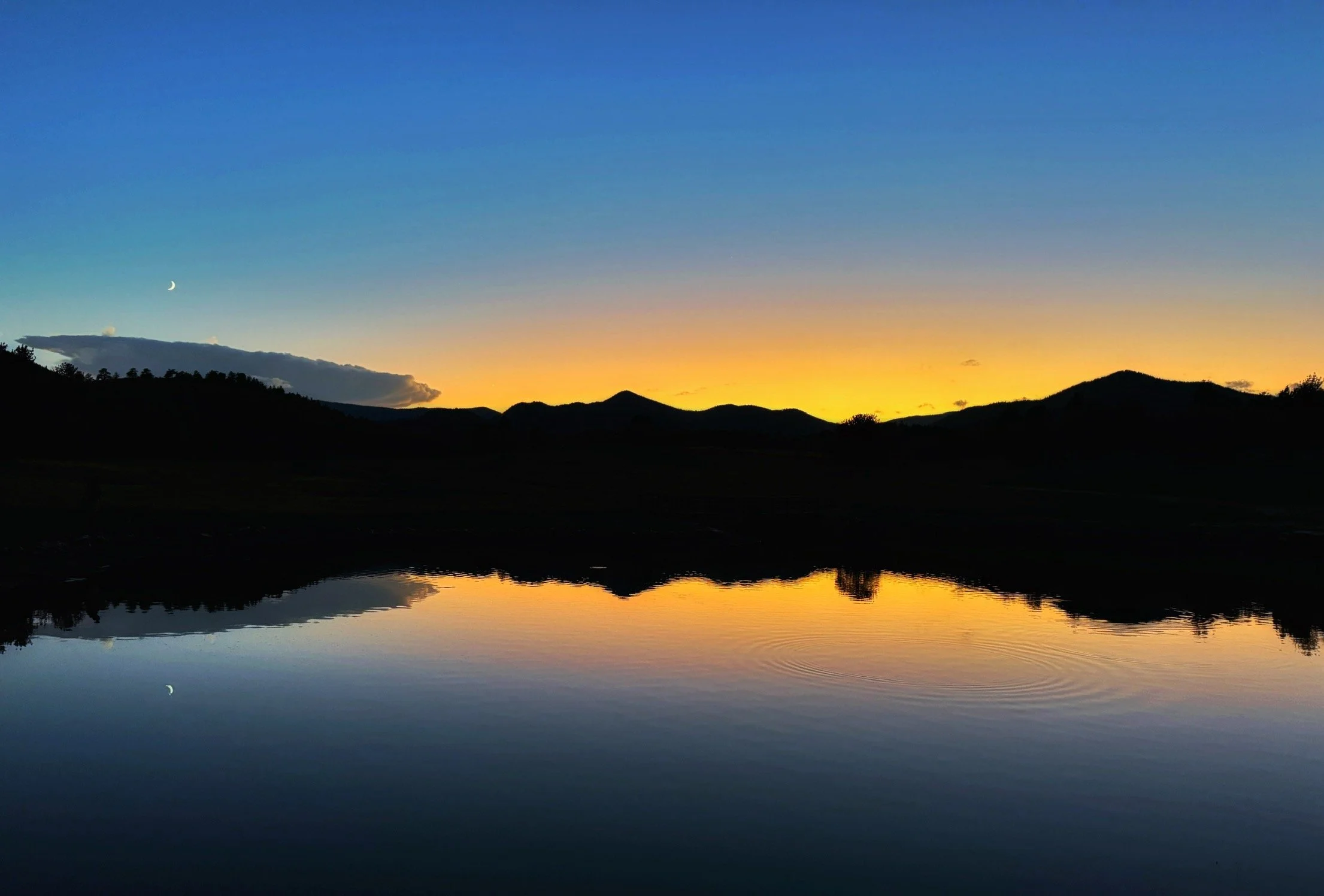 Silhouette of mountains and trees at sunset with a crescent moon in the sky, reflected in a calm body of water.