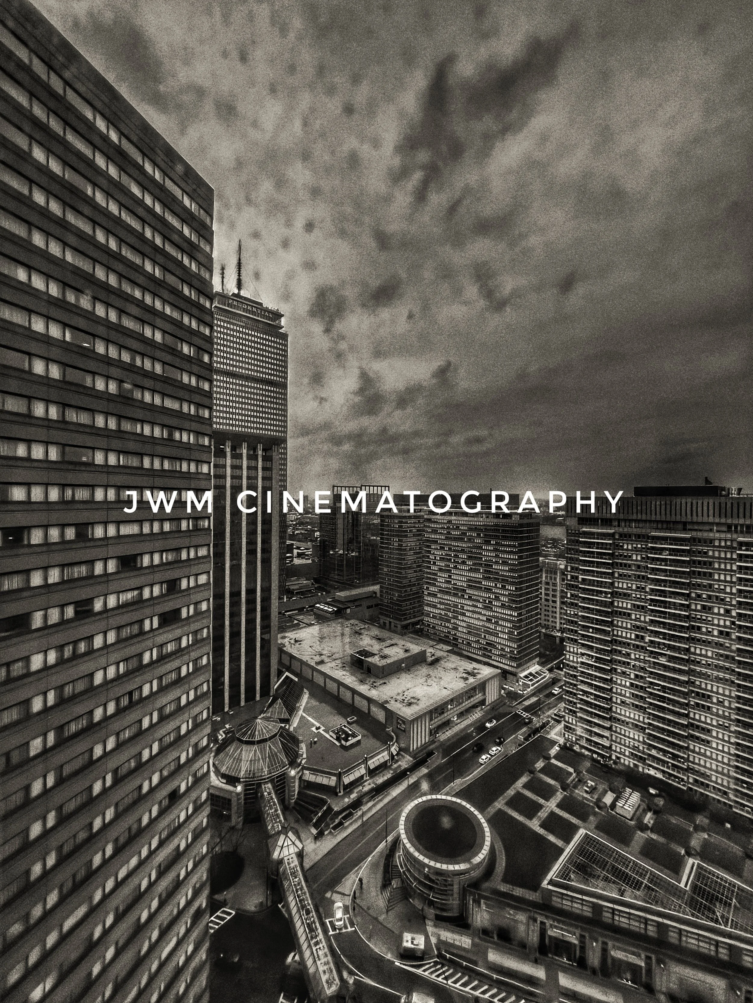 Aerial view of urban skyscrapers under cloudy skies, with a prominent building displaying an advertising sign and modern geometric architecture visible. Text overlay reads 'JWM Cinematography.'