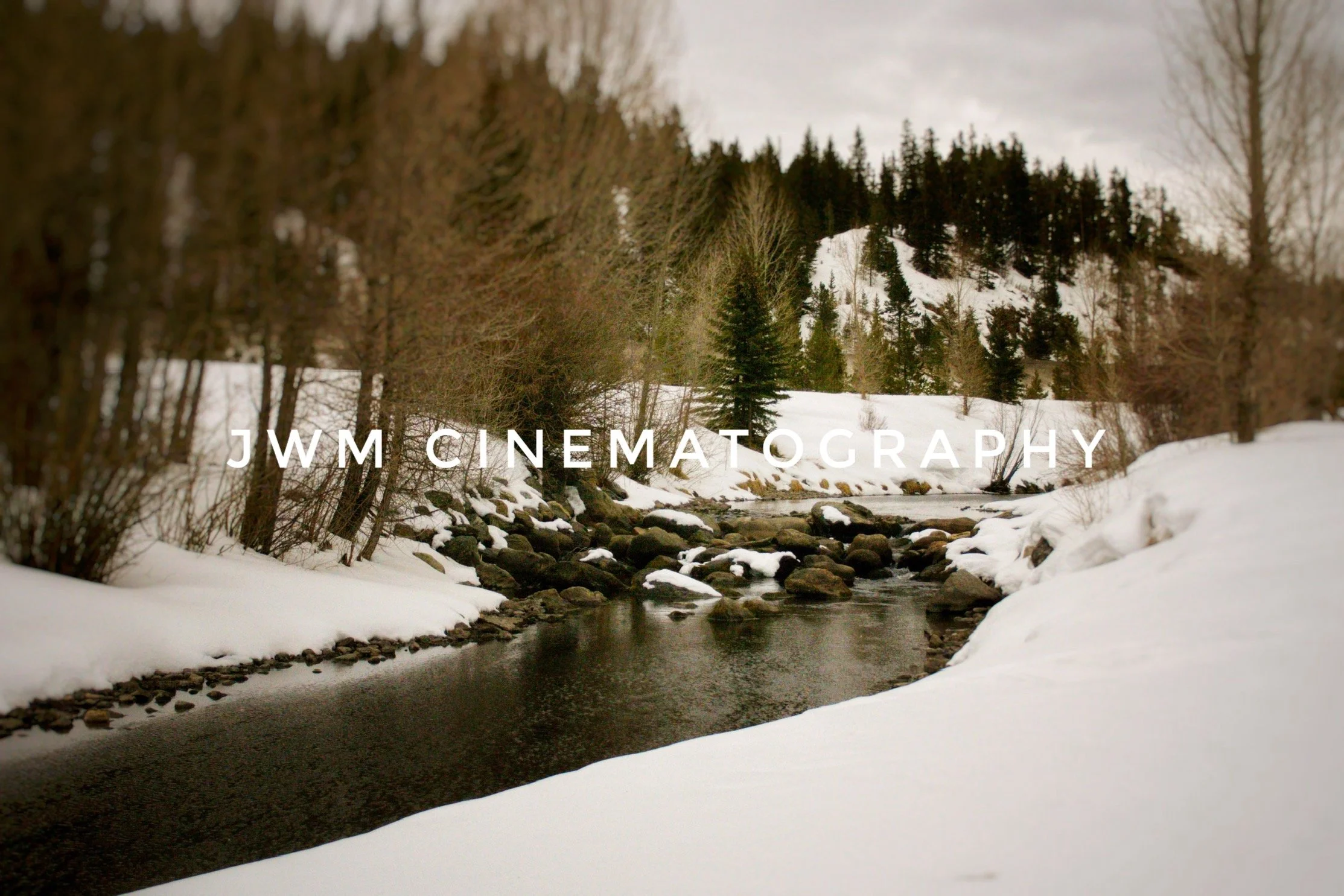 Winter landscape with a snow-covered stream and trees, featuring "JWM Cinematography" text overlay.