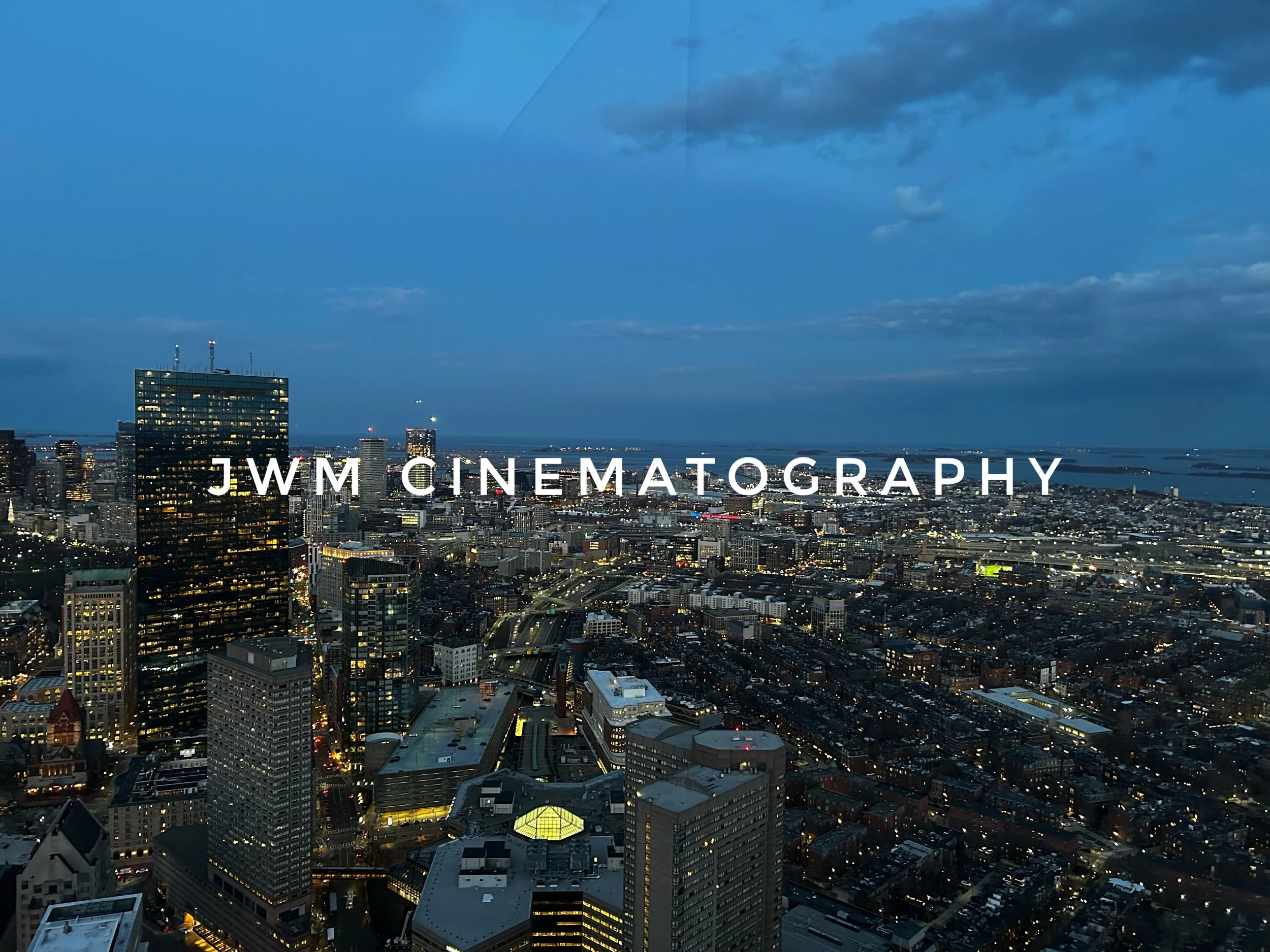 Aerial view of a city skyline at dusk with illuminated buildings and a water body in the background. Text "JWM Cinematography" is overlaid on the image.