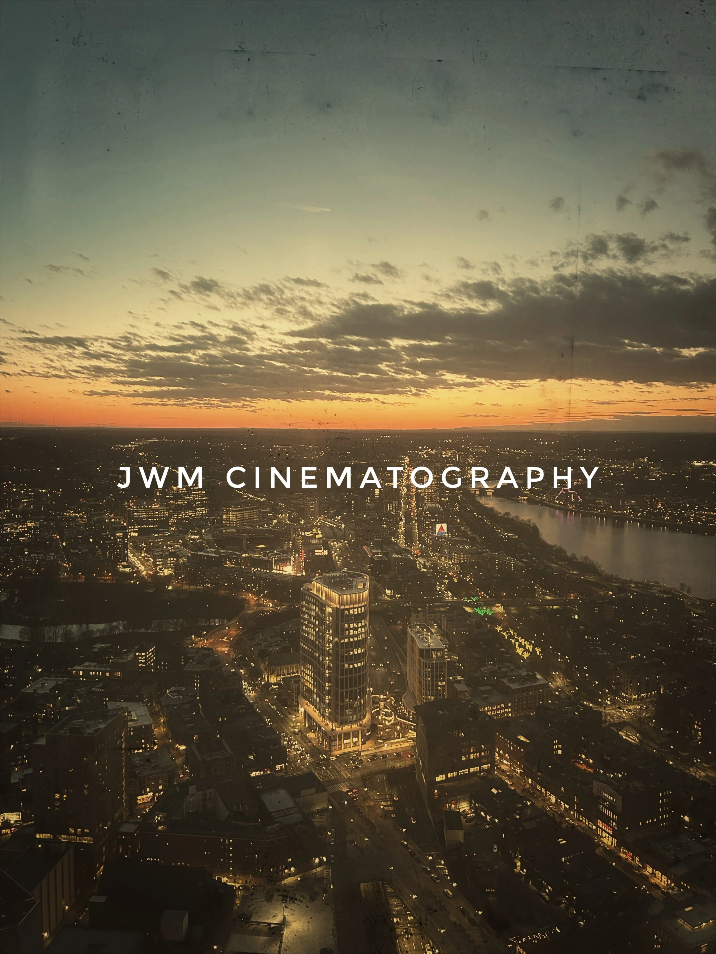 Aerial view of a cityscape at sunset with illuminated buildings, streets, and a river. Text overlay says 'JWM Cinematography.'