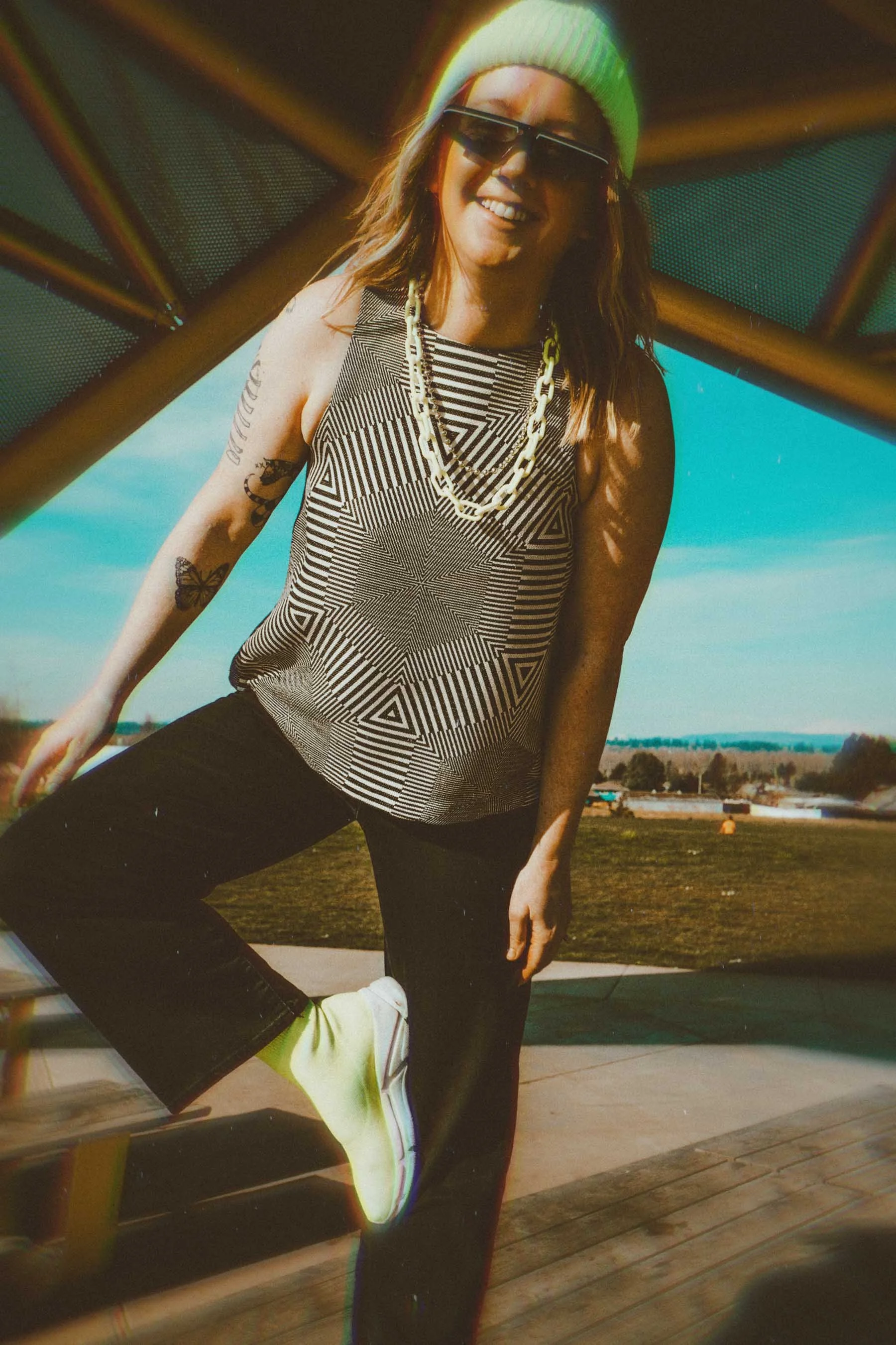 A woman wearing sunglasses, a white knit beanie, a patterned sleeveless top with geometric lines, black pants, and neon yellow sneakers, smiling outdoors on a sunny day.