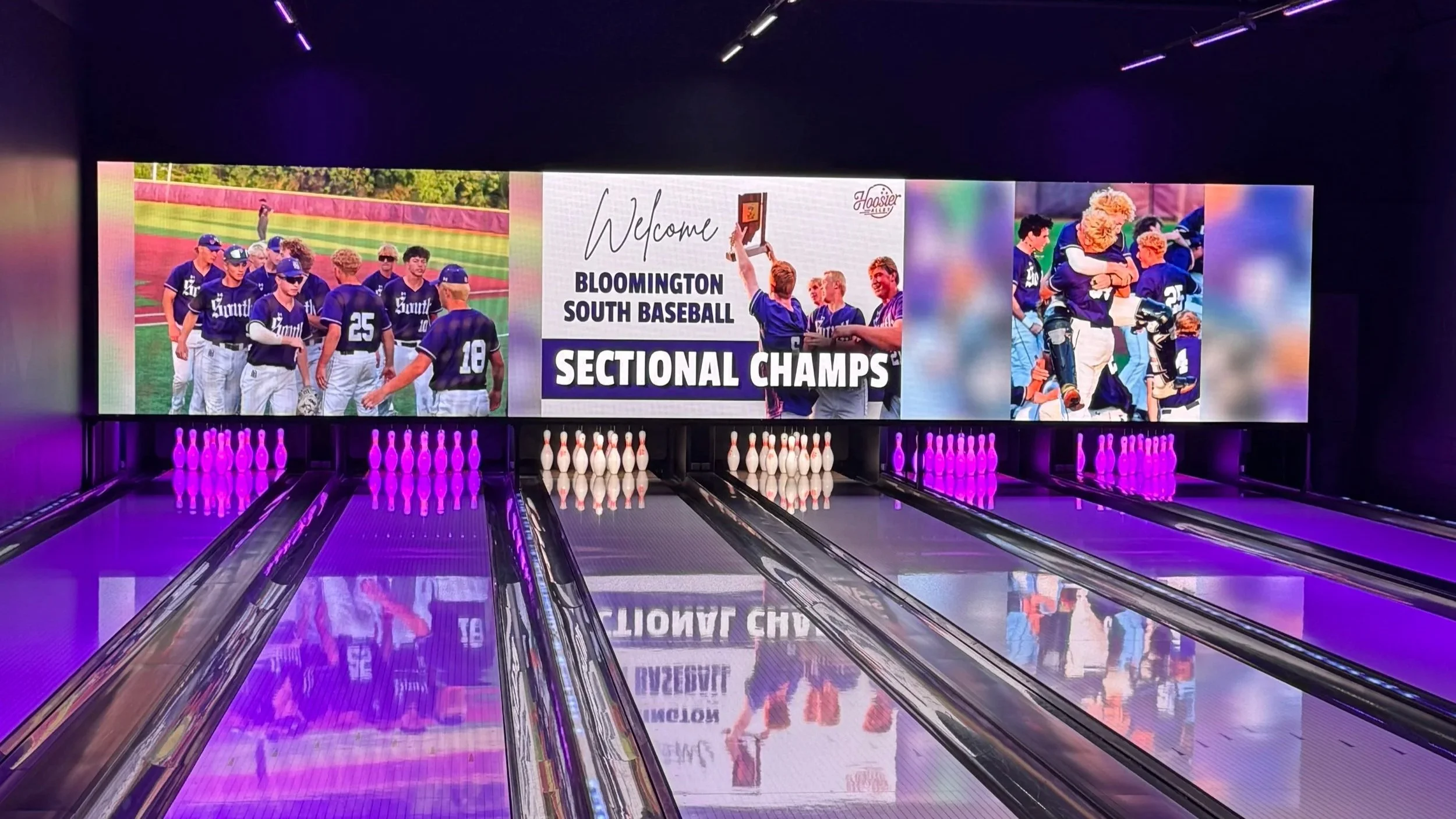 A bowling alley with a large screen displaying images of a baseball team, a welcome message for Bloomington South Baseball Sectional Champs, and people celebrating. Bowling pins are set up at the end of the lanes illuminated by purple lights.