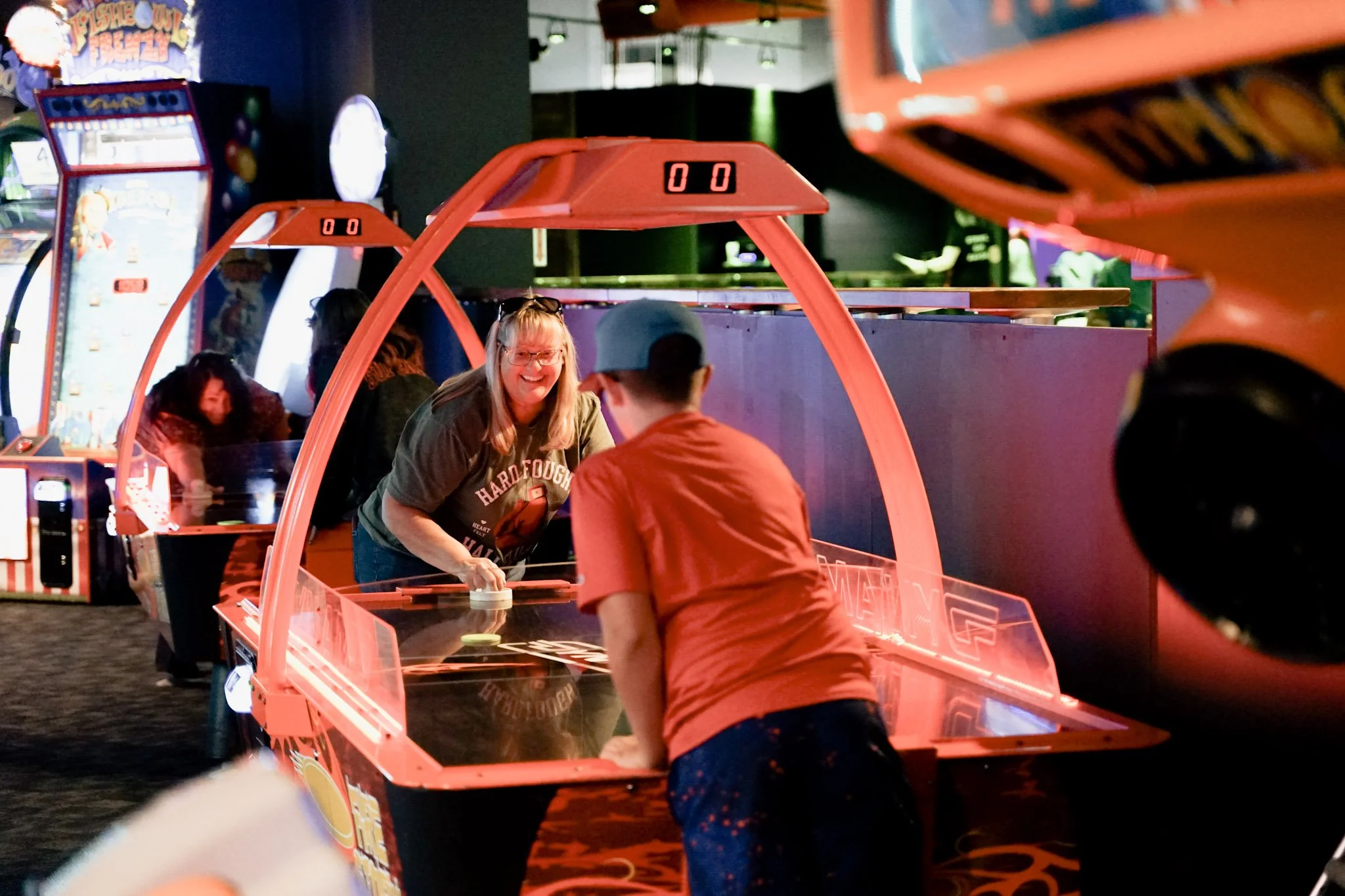 People playing air hockey at an arcade, with colorful arcade machines in the background.