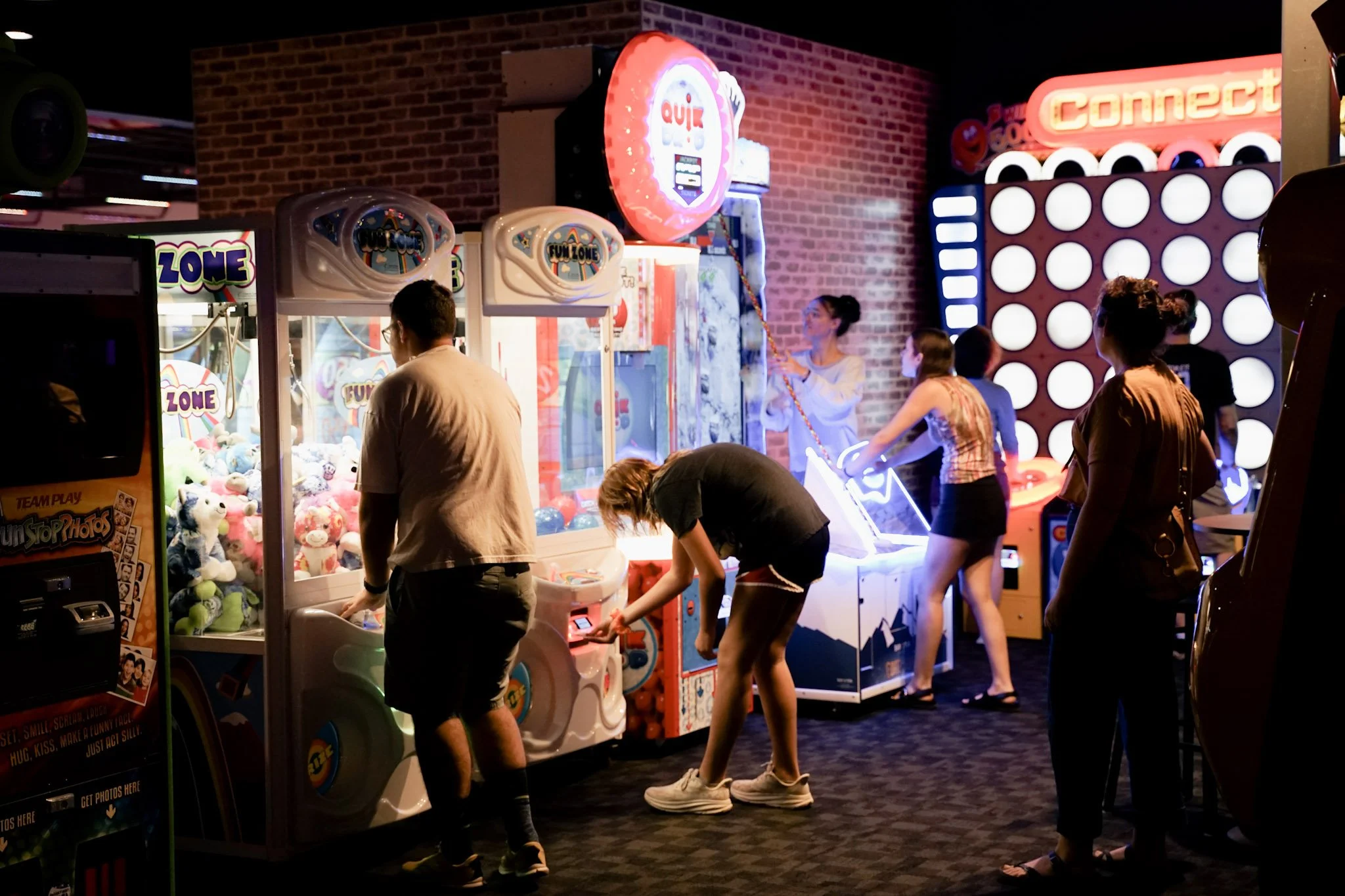 People playing arcade claw machine games at an arcade with colorful neon signs and brick wall decor.