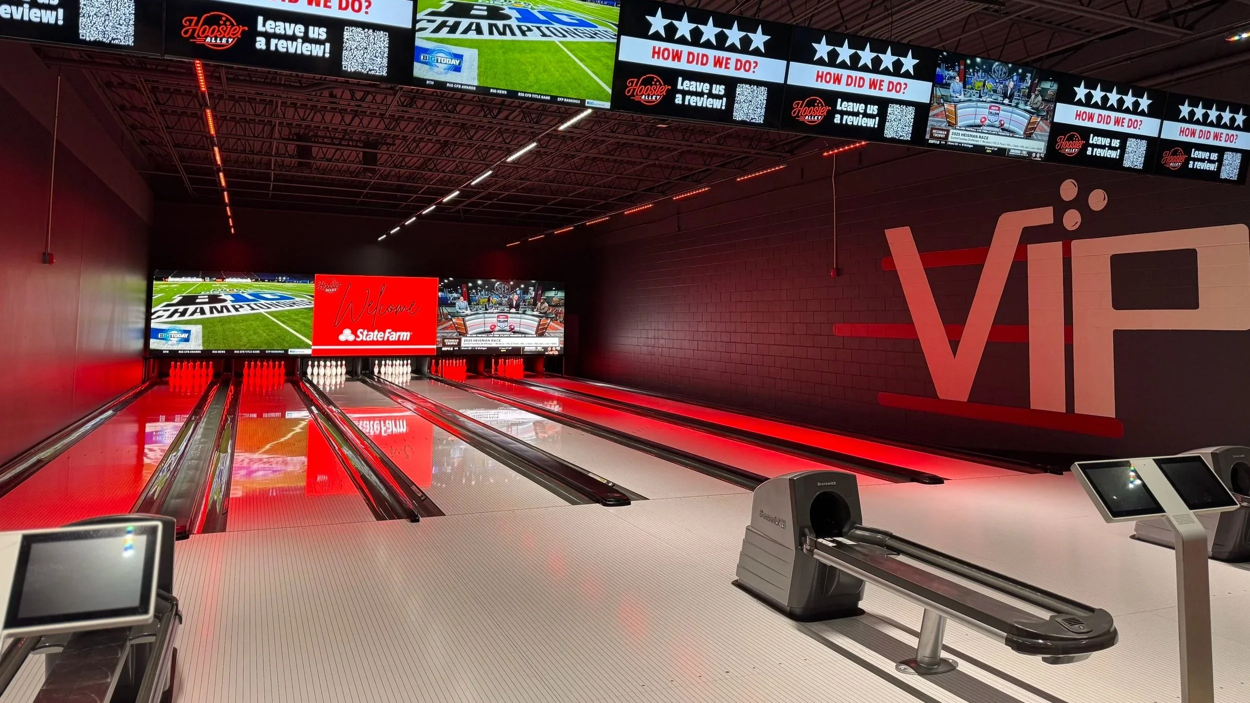 Indoor bowling alley with lanes, electronic scoring monitors, and a dark wall with large Red and White Hoosier Alley signage. Multiple advertisement screens are mounted above the lanes.