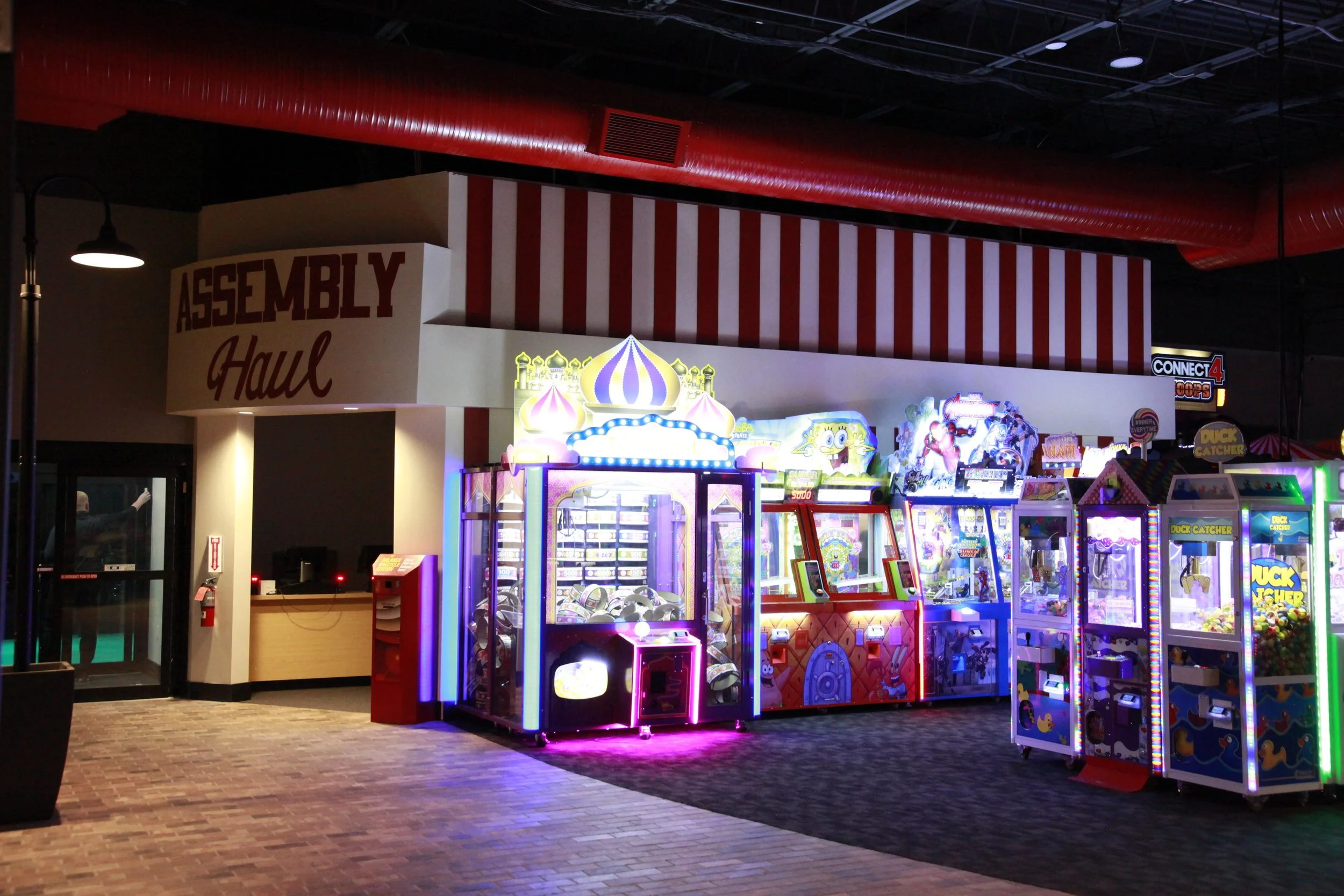 Inside an arcade with bright, colorful game machines, and a sign that says 'Assembly Haul' in red letters on a white background, with red and white striped decorations and a ceiling with exposed pipes.