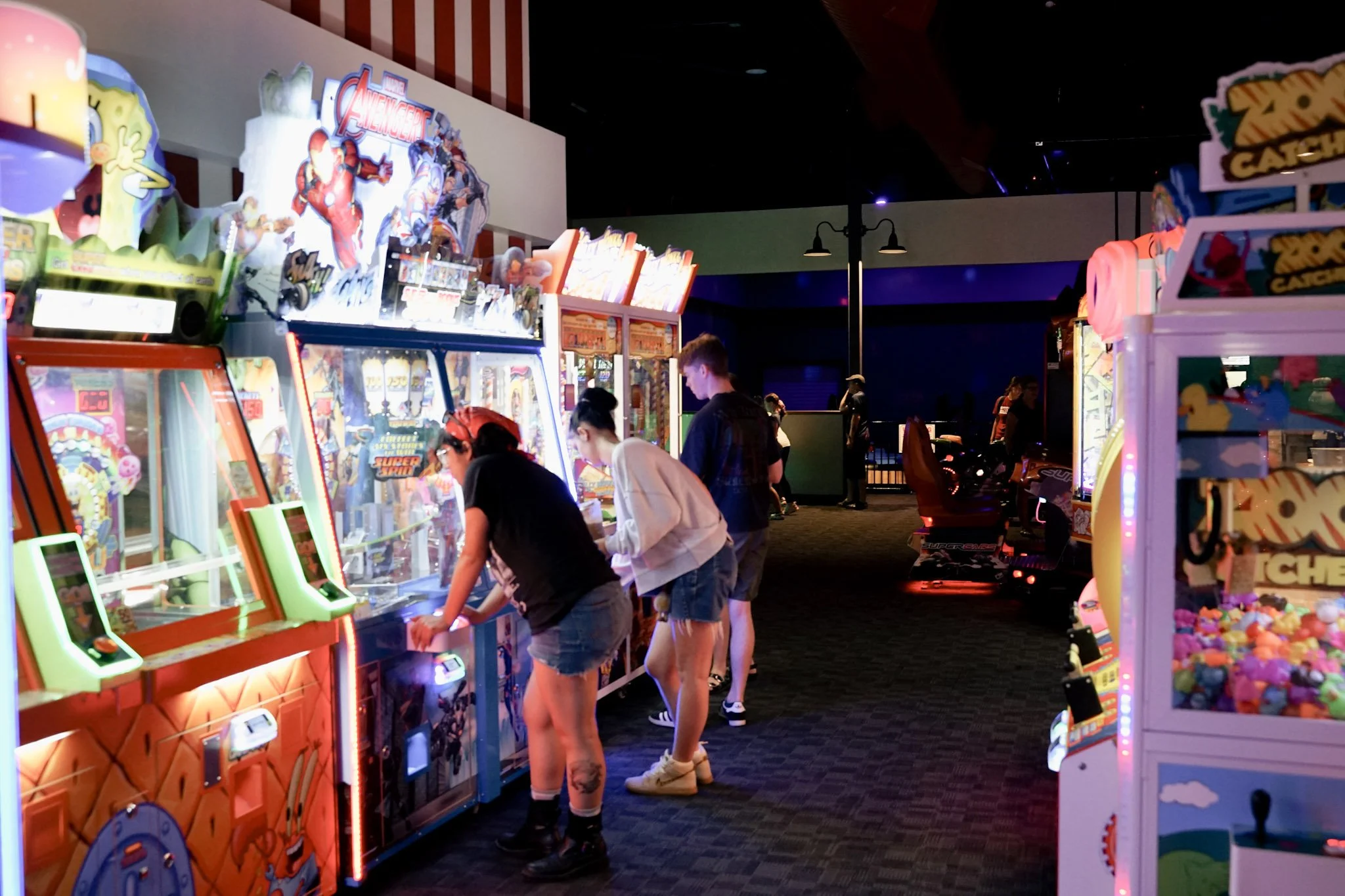 People playing arcade games in a brightly lit arcade with colorful machines and a dark ceiling.