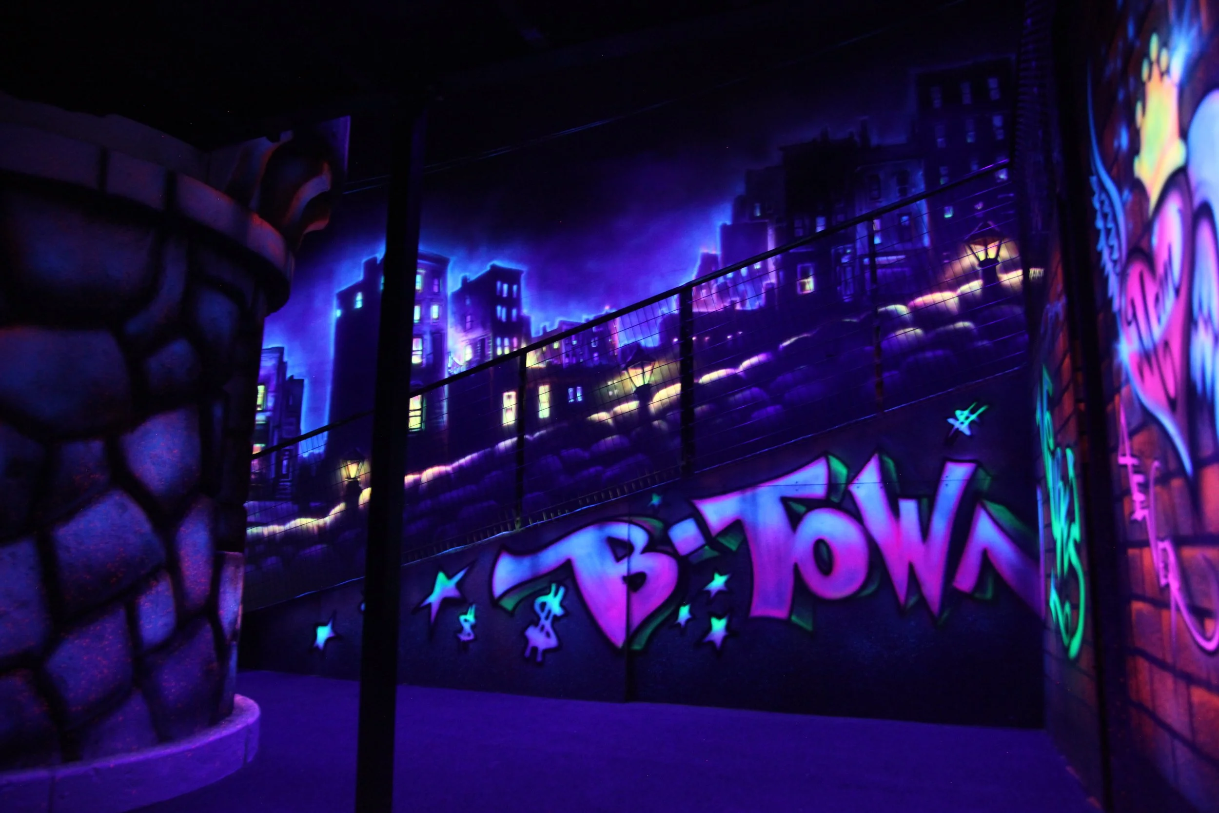 Brightly colored graffiti art on a wall featuring stars and playful lettering, illuminated by blacklights with a cityscape mural background and a section of a chain-link fence in the foreground.