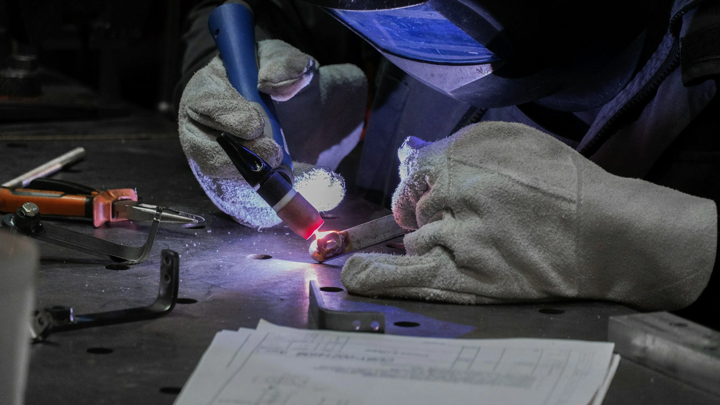 Close-up of a welder using a TIG welding torch on a metal piece, wearing protective gloves and a welding helmet, with tools and blueprints on the table.