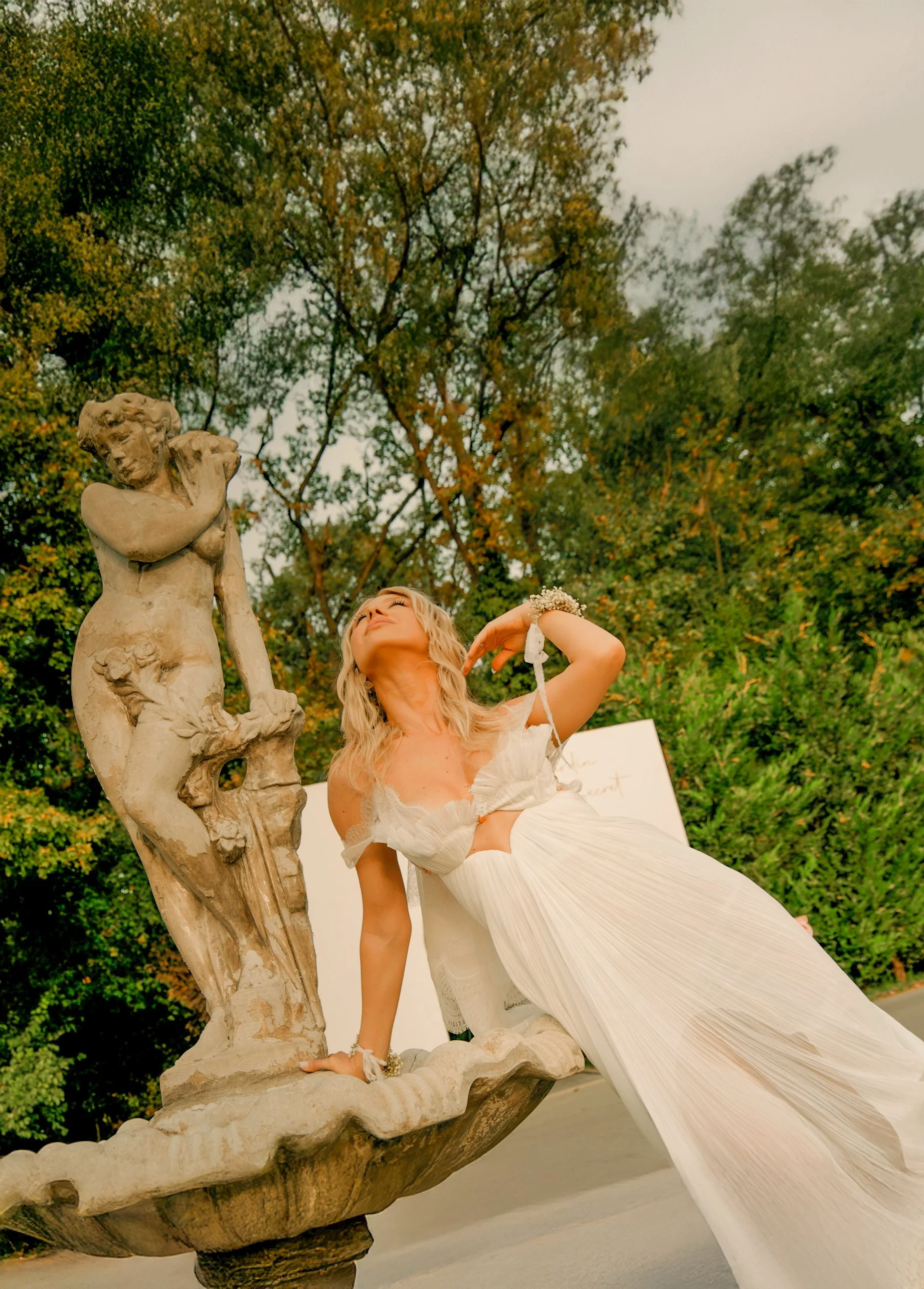 Woman in a white dress posing next to a stone sculpture outdoors with trees in the background.