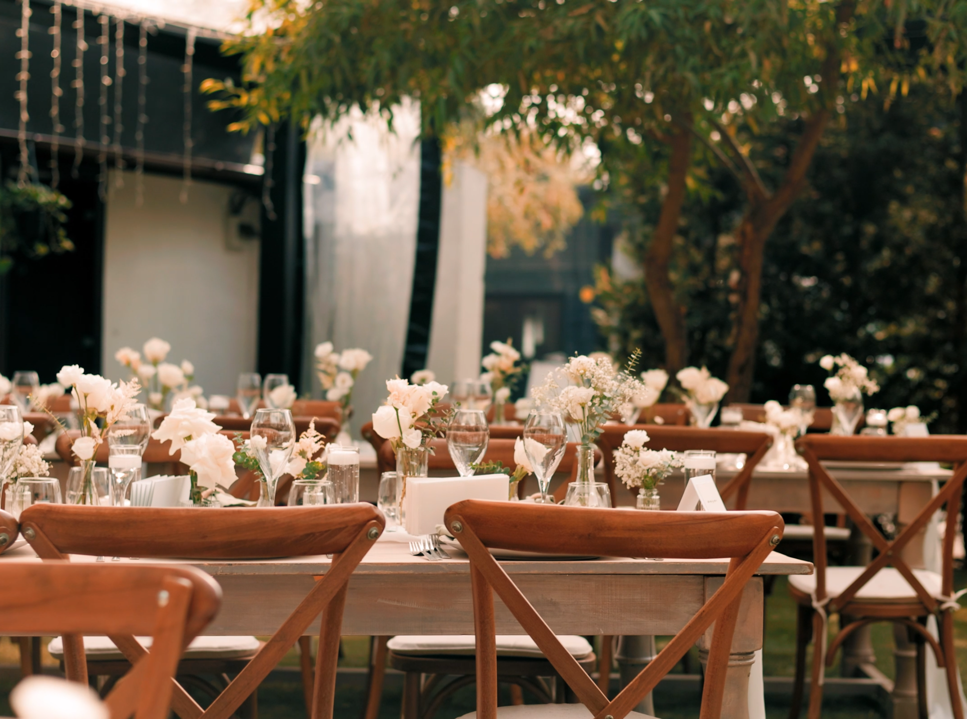 Outdoor wedding or event reception table set with white flowers, glassware, and silverware in a garden or yard with trees and string lights.