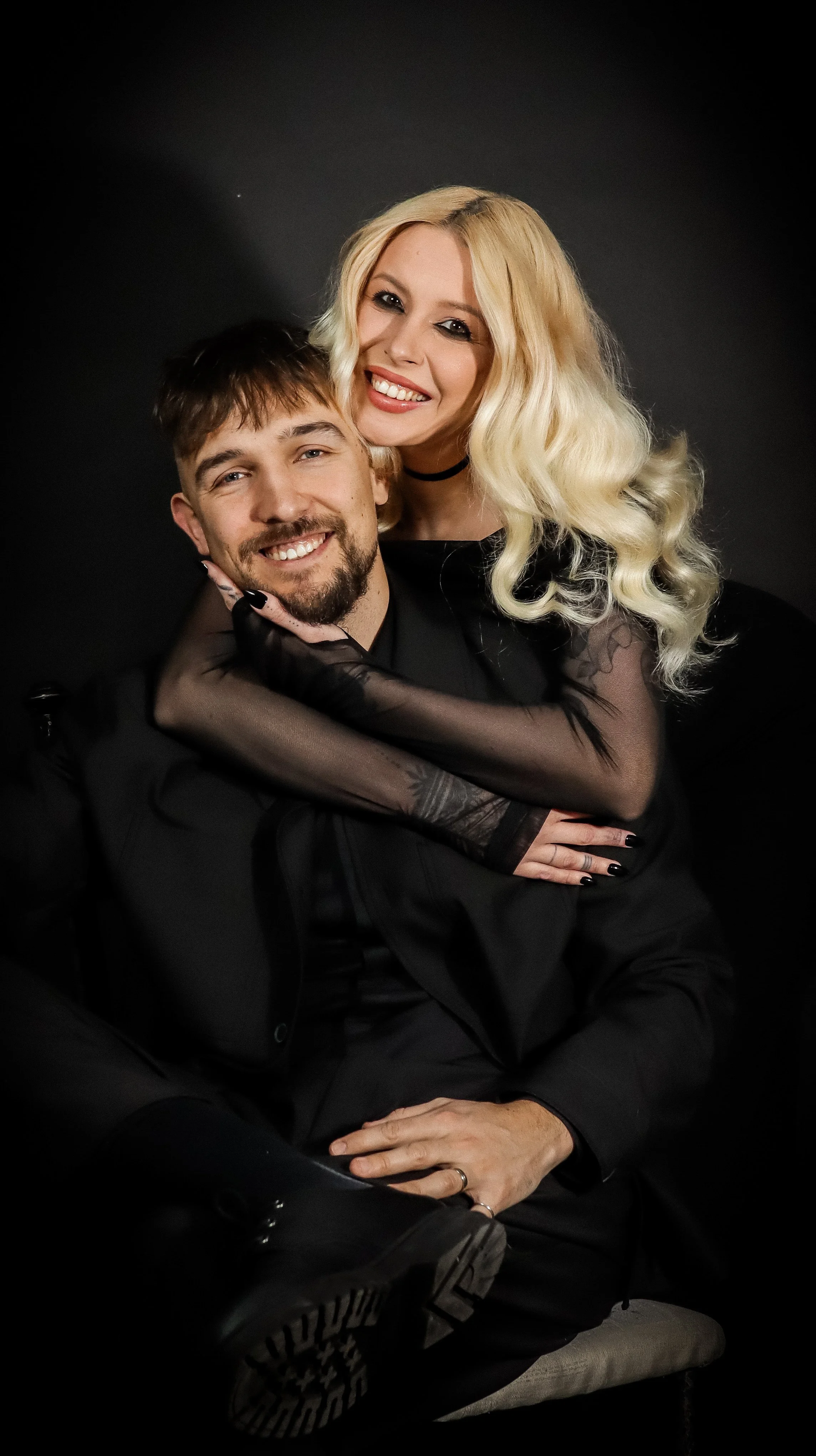 A smiling couple dressed in black, with the woman embracing the man from behind, against a dark background.