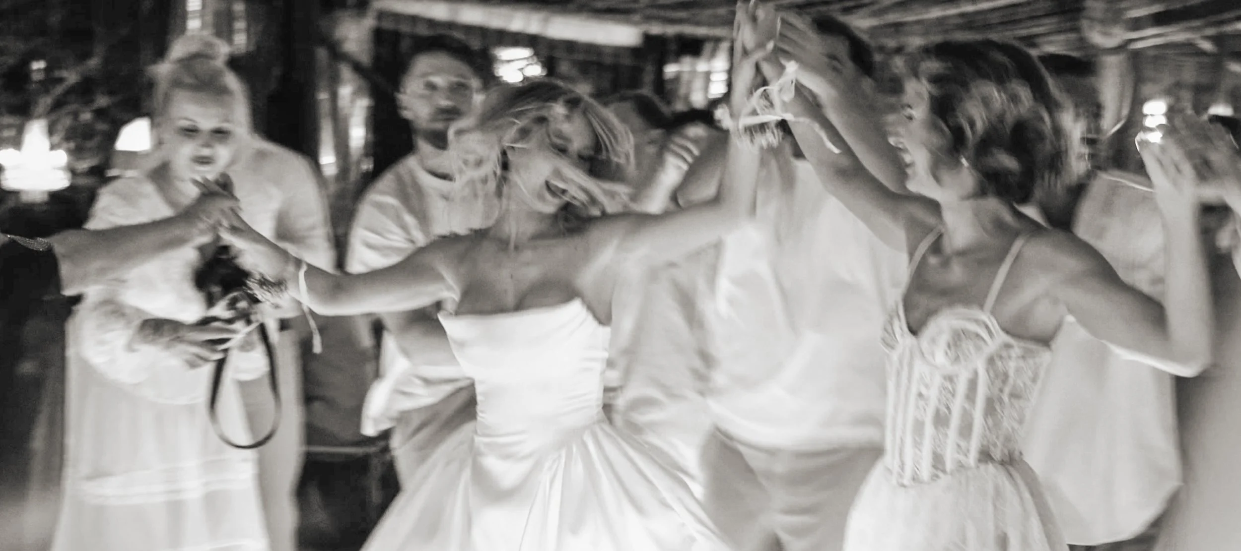 Women dancing and celebrating at a wedding reception, wearing white dresses, in a festive indoor setting.