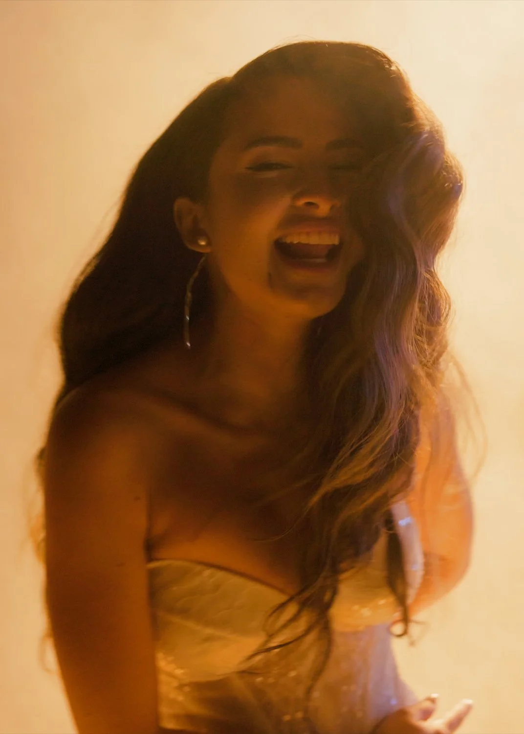 A woman with long, wavy hair is smiling and laughing, wearing a strapless dress, with warm lighting and a plain background.