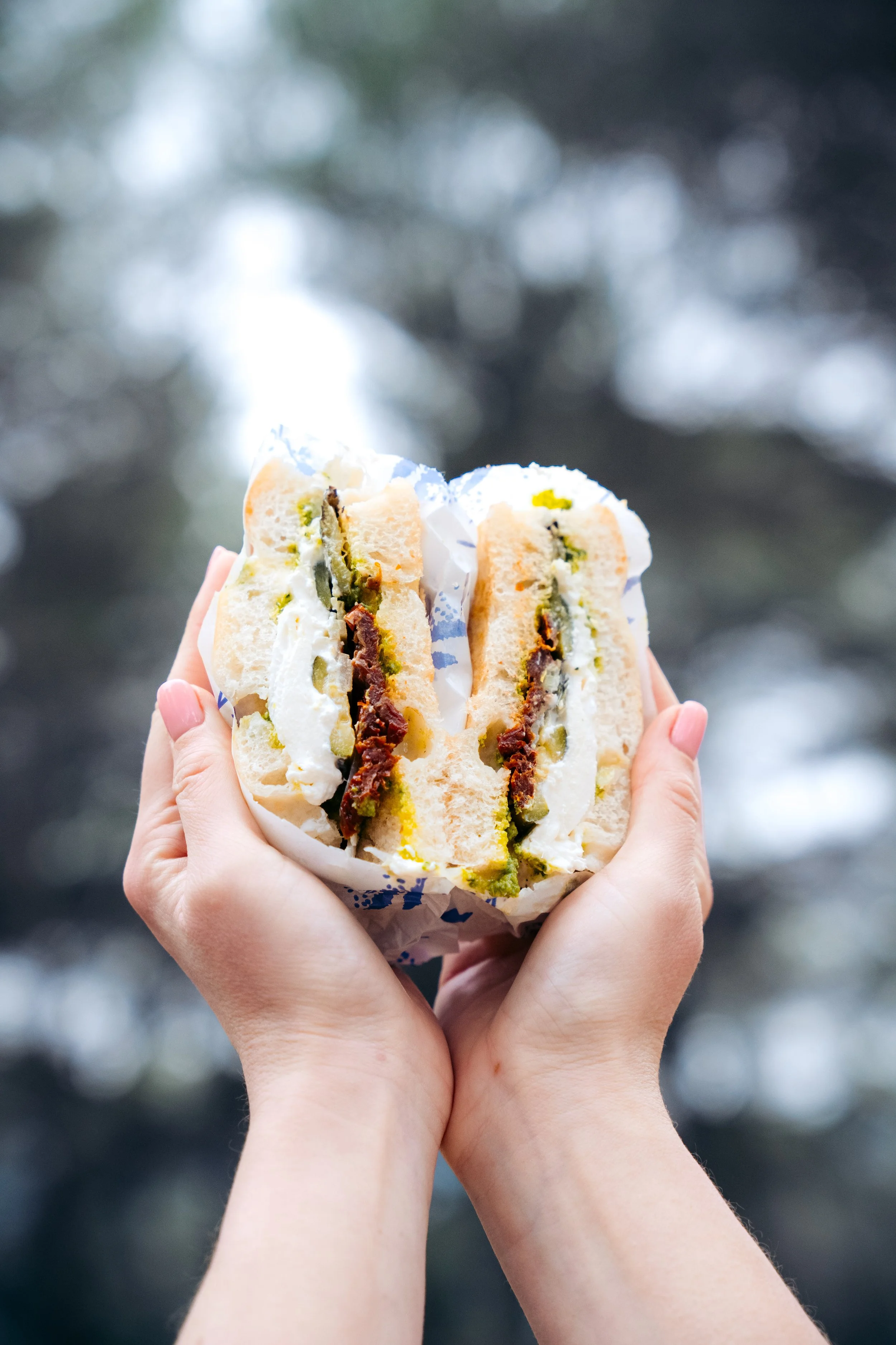 Deux mains tiennent un sandwich garni de fromage, tomates séchées, et pesto, enveloppé dans du papier.