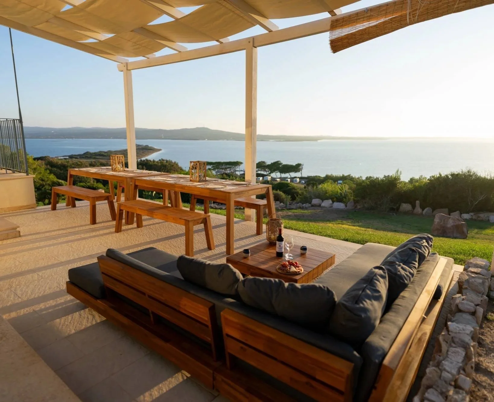 Zona living con divano e tavolo vista lago, con tavolo e panche in legno, decorazioni e bottiglie di vino, sotto un pergolato, con paesaggio naturale e acqua sullo sfondo.