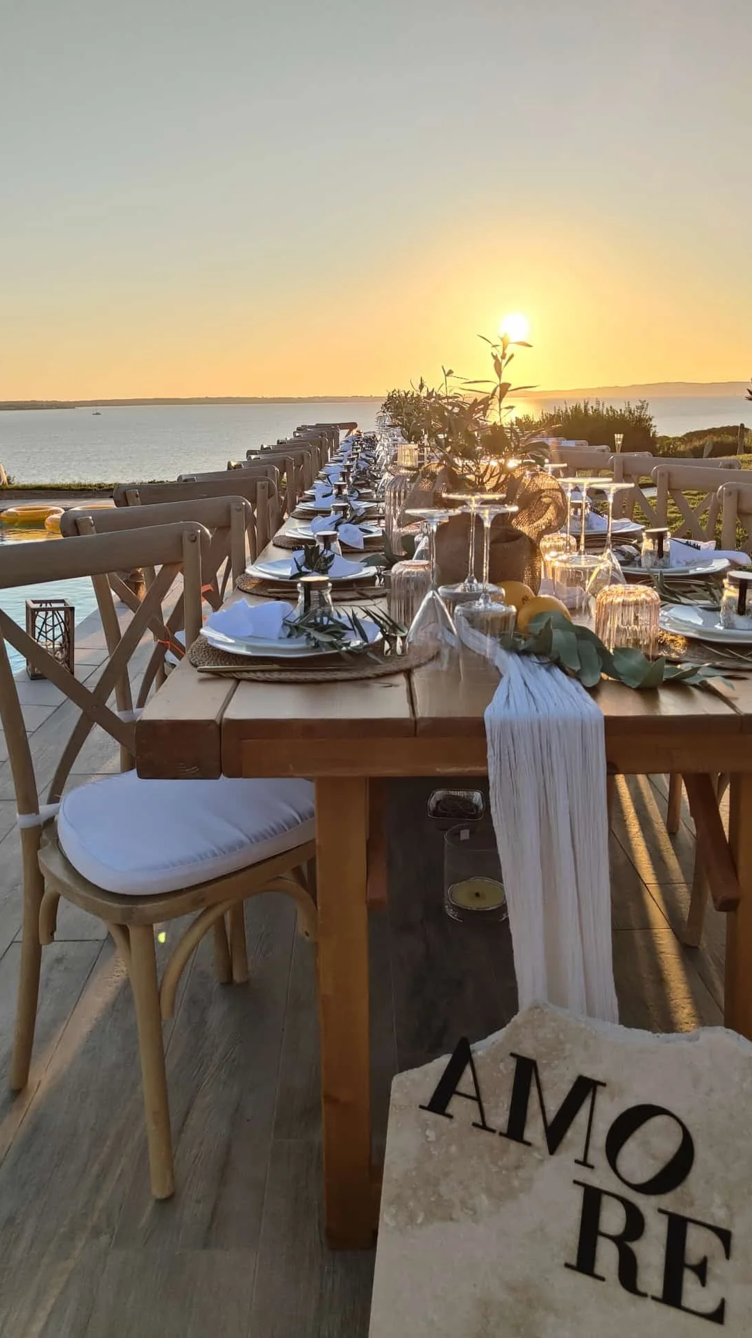 matrimonio in sardegna con Tavolo apparecchiato per una cena all'aperto con vista sul mare al tramonto, decorato con luci e candele, con un cartello con la scritta 'AMORE' appoggiato sul pavimento.