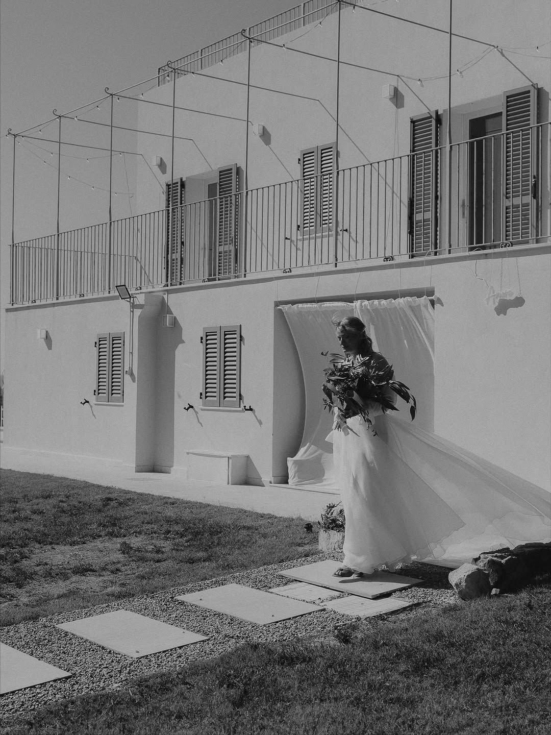 matrimonio in Sardegna con Donna in abito da sposa con bouquet di fiori, fuori da una casa bianca con finestre con persiane, sotto un cielo chiaro.