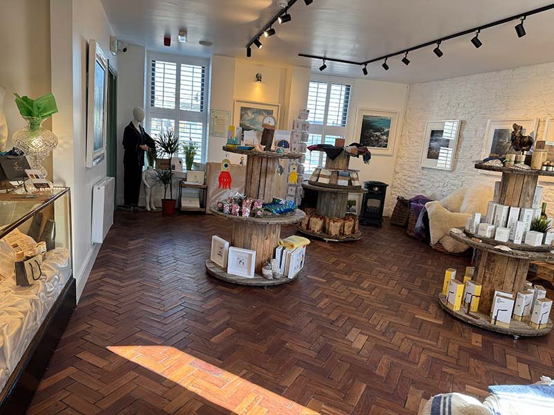 Interior of a boutique shop with wooden display tables, art on the walls, a white brick wall, sunlight coming through large windows, and various home decor items.