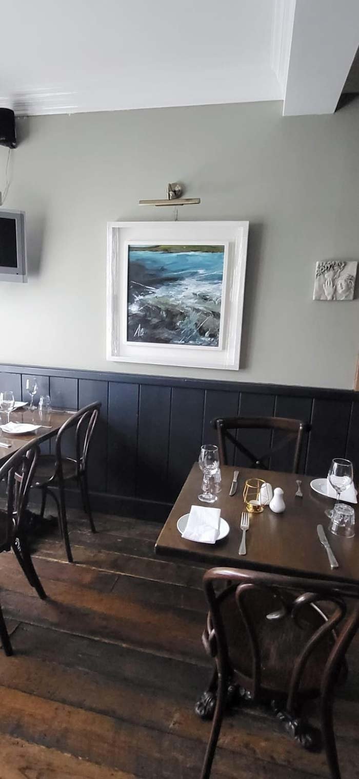 Dining area with dark wood tables and chairs, glassware, plates, and silverware set for multiple guests. A framed painting of a body of water hangs on a light-colored wall, and a small TV is mounted on the wall. The floor is wooden planks, and there