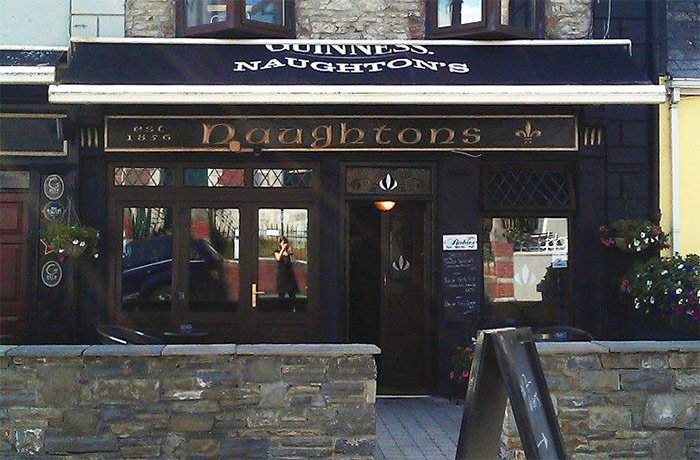 Front view of Naughton's pub with black exterior, stone foundation, and flower pots on both sides of the entrance