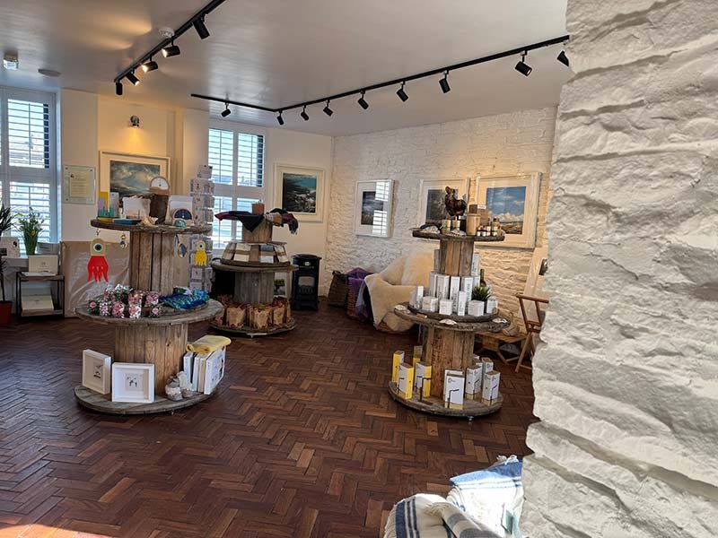 Interior view of an art gallery or gift shop with framed paintings on white brick walls, wooden display tables with various art pieces and gifts, and a sitting area with a cream armchair. Large windows with plantation shutters let in natural light, a