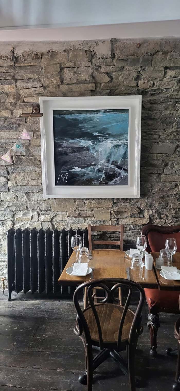 Interior of a restaurant with a rustic stone wall, a framed painting of a seascape, a black radiator, and a wooden table set with glasses, plates, and utensils.