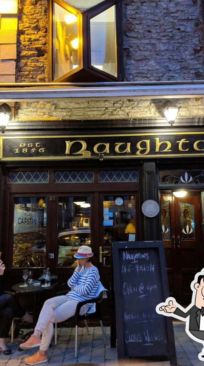 People sitting outside a pub called 'Naughtons' established in 1856, with a woman wearing a striped shirt and a pink hat. The pub has a black and gold sign, large windows, and a chalkboard sign displaying open hours and contact information.