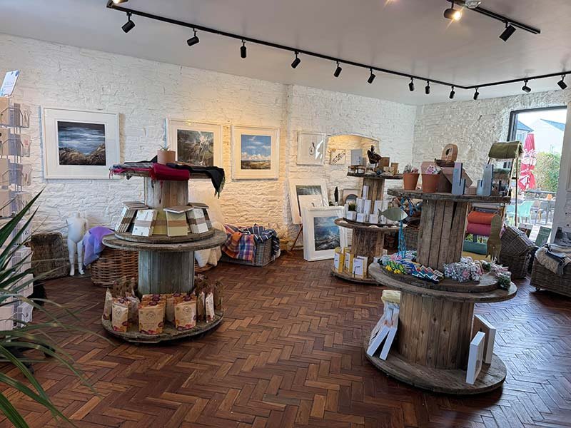 Interior of an art and gift shop with framed landscape photos on white brick walls, wooden tables displaying books, art supplies, and decorative items, and hardwood flooring.