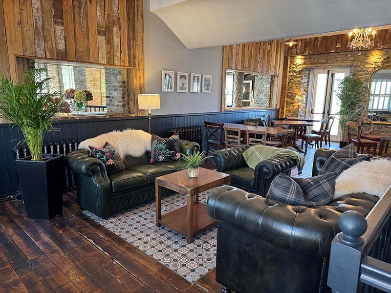 Cozy living room with black leather sofas, wooden coffee table, plants, and decorative pillows, featuring wood-paneled walls and a mix of rustic and modern decor.