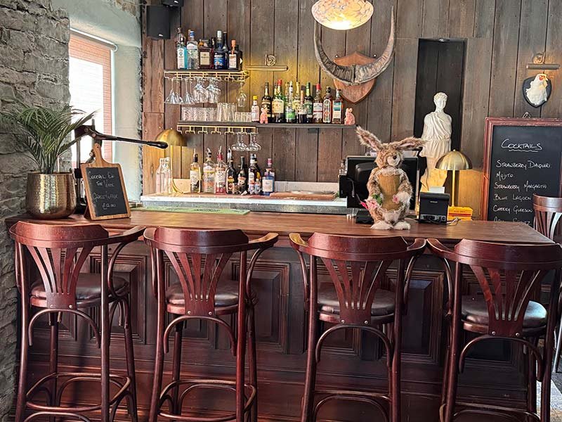 A cozy bar with a wooden counter and five matching wooden barstools. Behind the bar, there are shelves with various bottles of liquor, glasses, and a chalkboard menu. There is a rabbit dressed in a costume sitting on the bar counter, and the backgrou