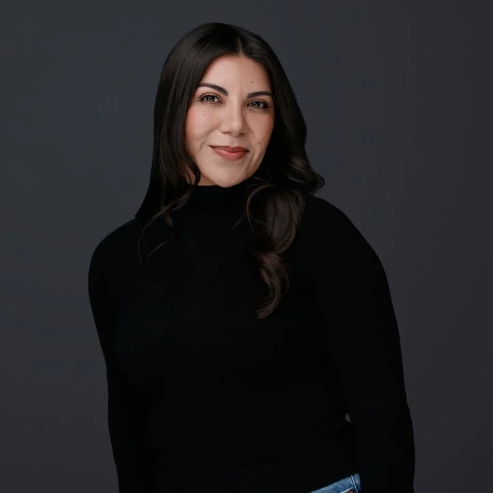 A woman with long, dark hair wearing a black turtleneck sweater, smiling slightly, against a dark gray background.