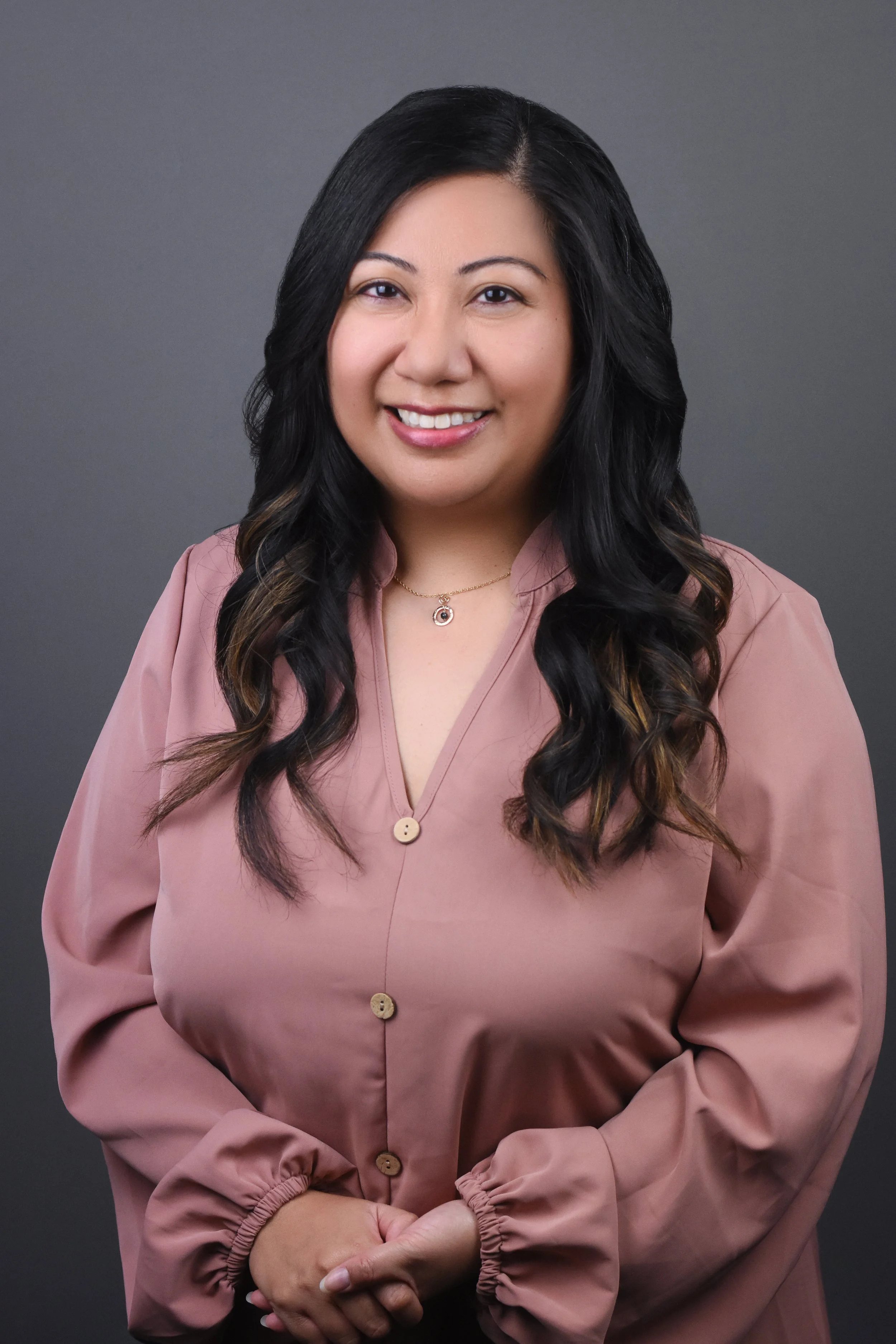 A woman with long black hair styled in loose waves, wearing a pink blouse with large buttons and a delicate necklace, standing against a gray background, smiling at the camera.