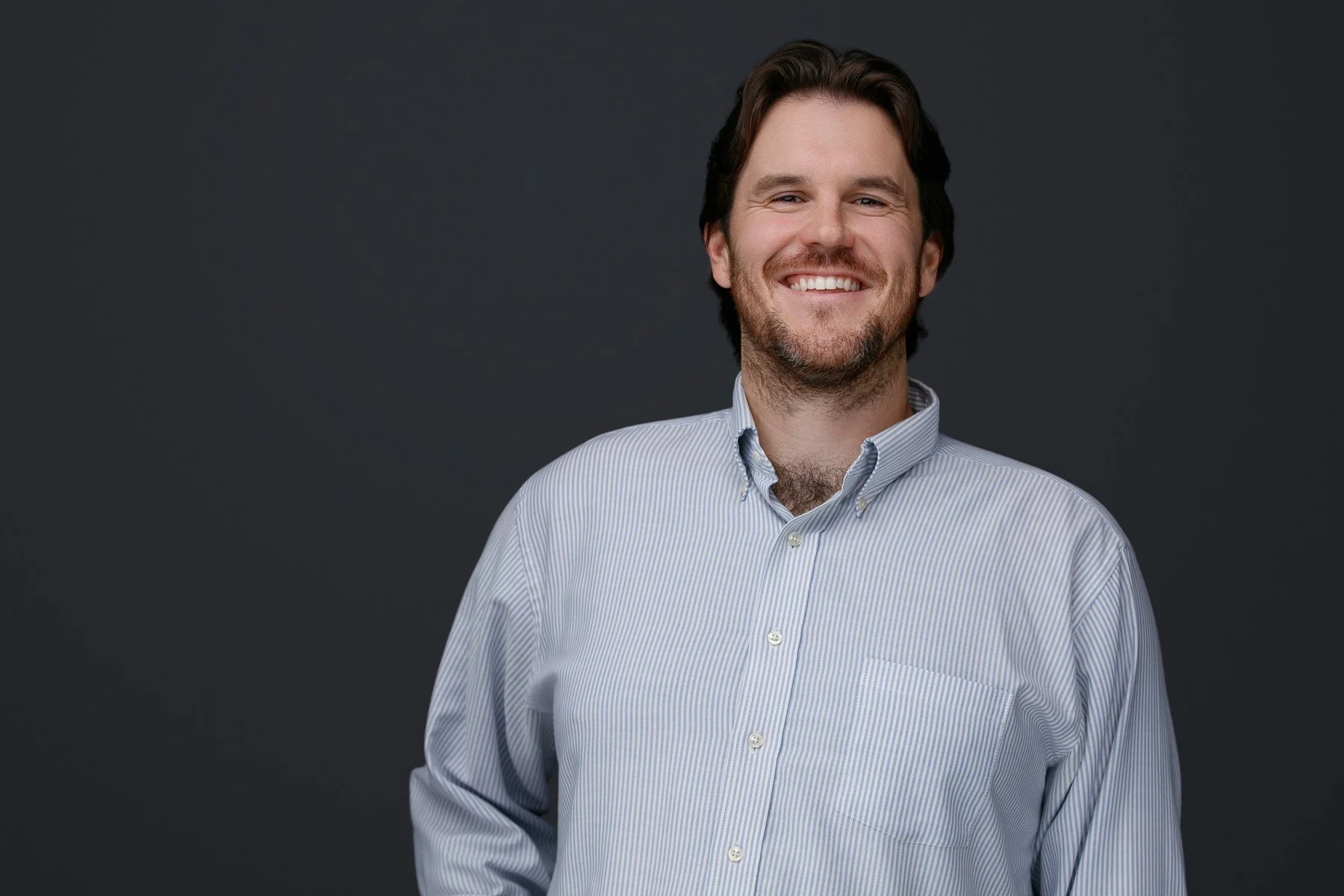 A smiling man with dark hair and a beard wearing a light blue striped button-up shirt, standing against a dark gray background.