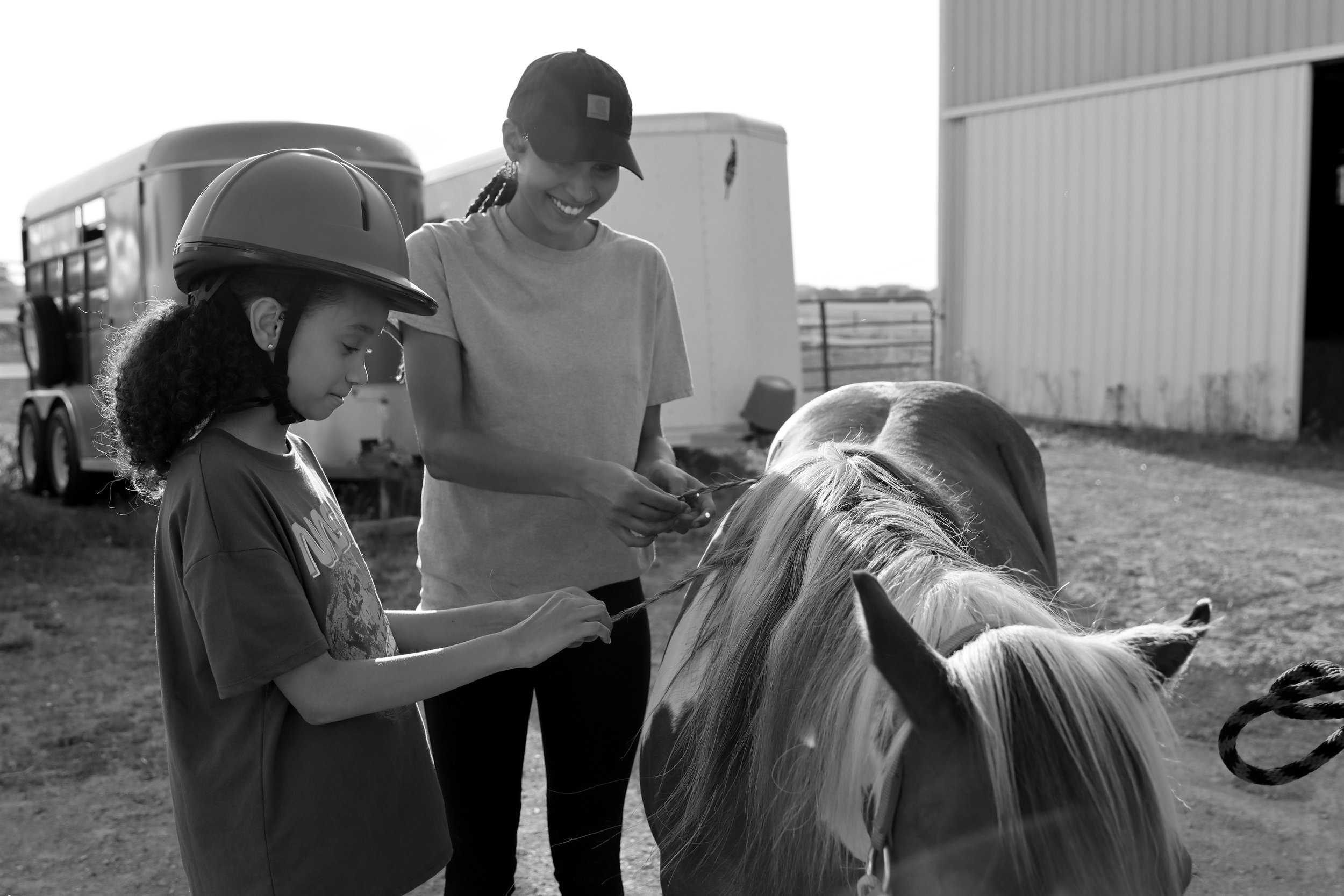A child wearing a helmet and a smiling adult are braiding a horse's mane outside near a stable and horse trailer.