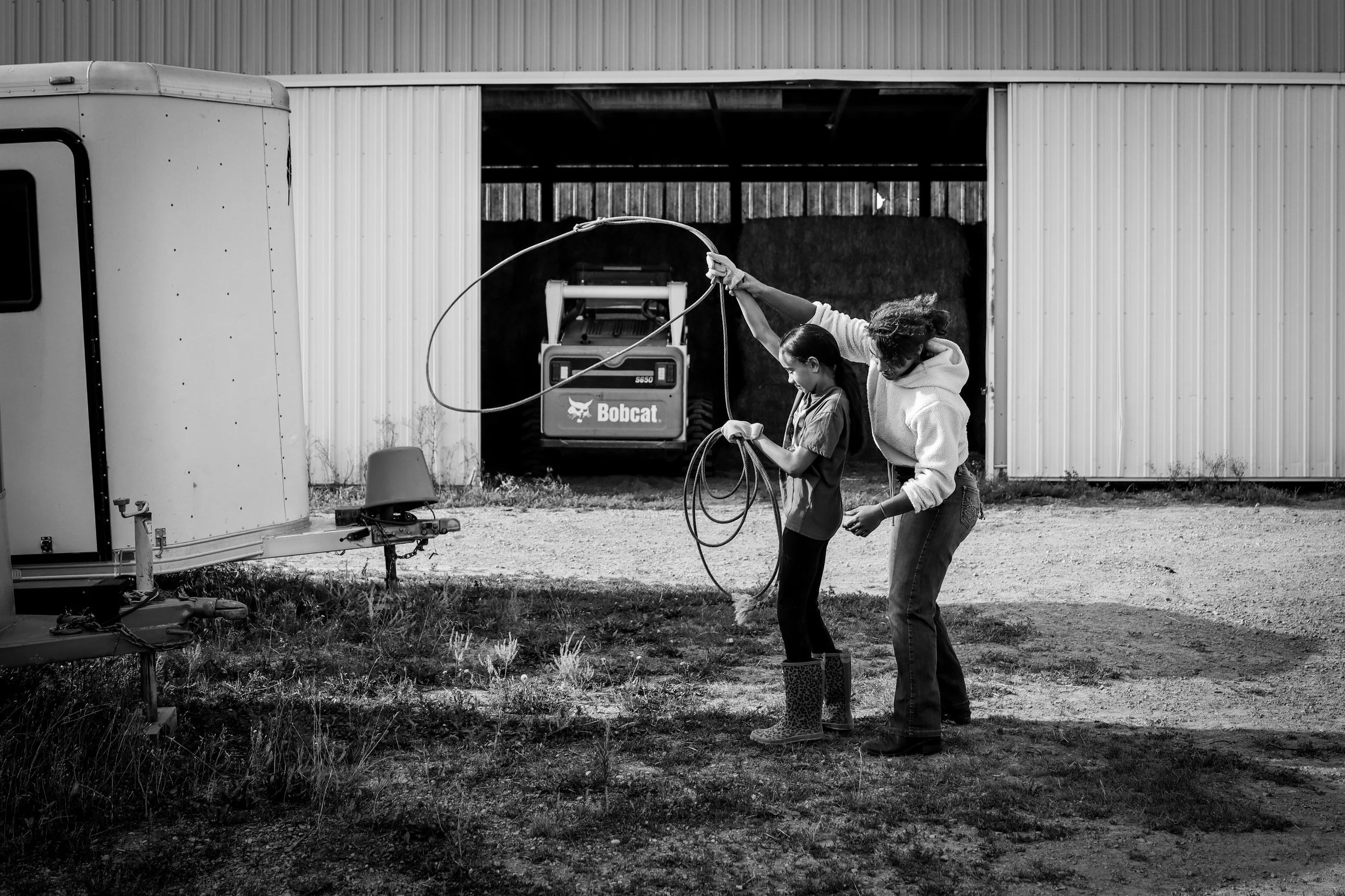 Two individuals practicing roping near a barn with a Bobcat vehicle visible inside, trailer hitch in the foreground.
