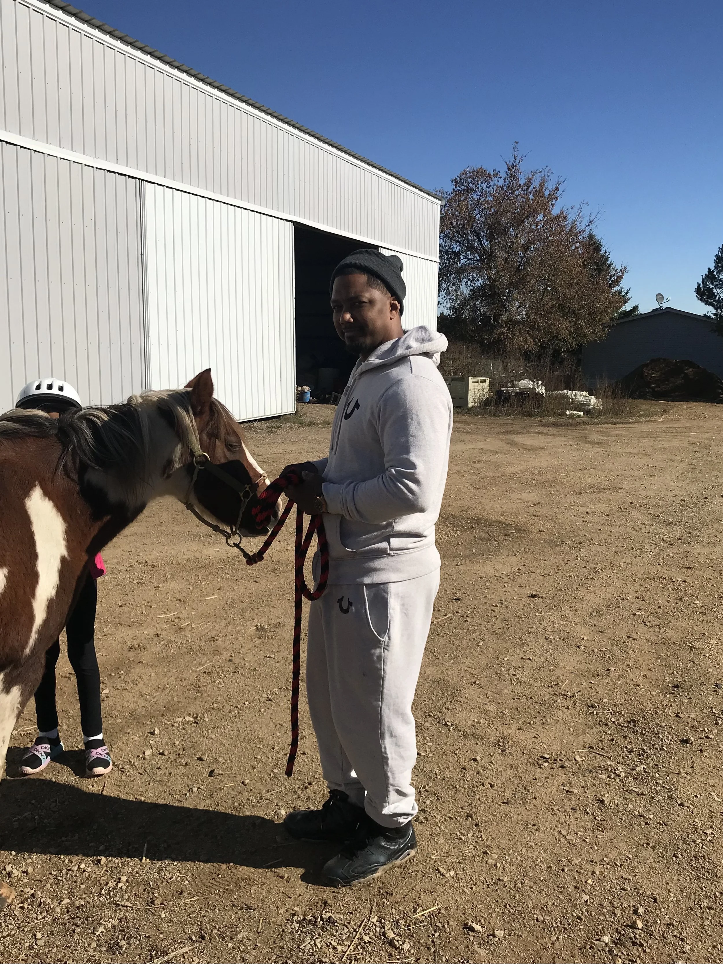 A man in a gray tracksuit and beanie holding a brown and white horse by the reins, with a person in a riding helmet partially visible. They are outside near a metal barn on a clear day.