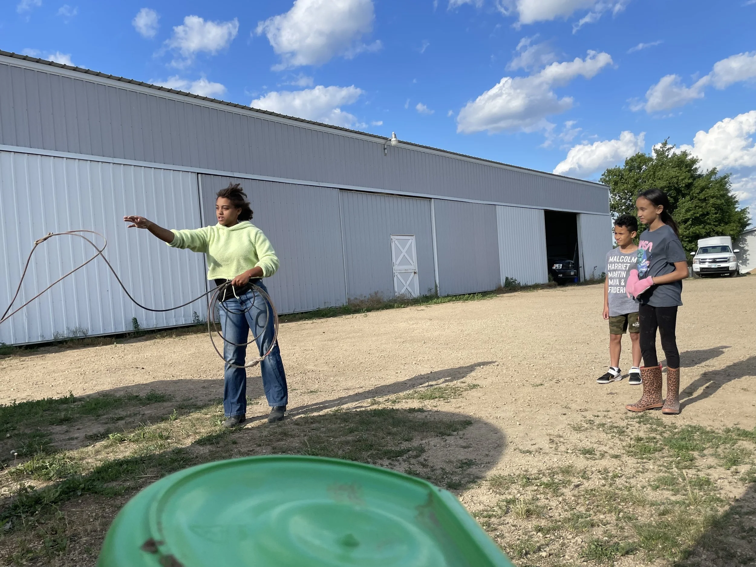 Three children outside a metal building, one using a rope lasso while the other two watch. The setting is a dirt and grass area with a clear blue sky.