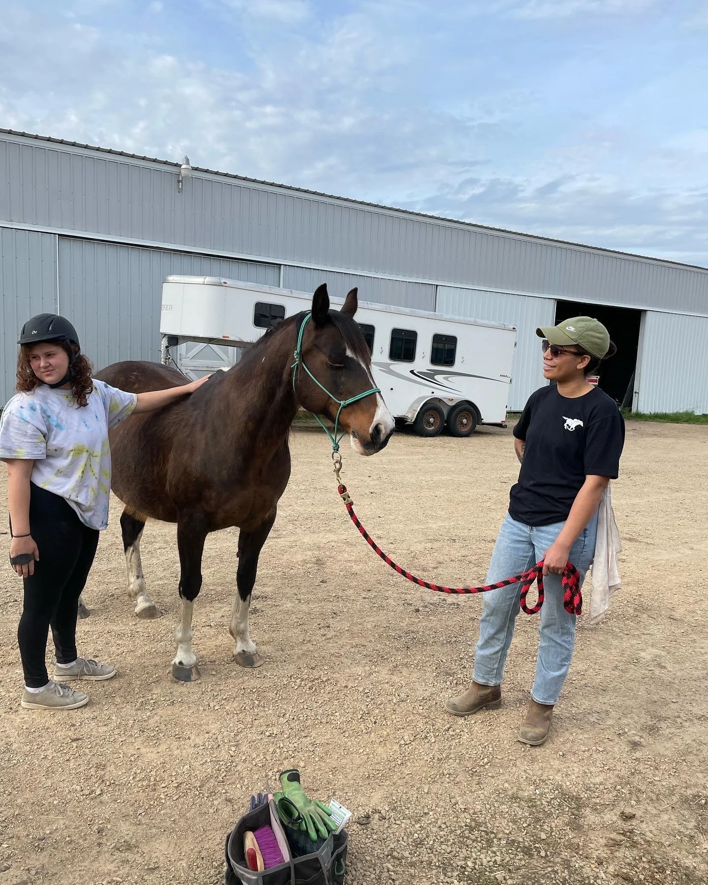 Two people with a horse near a barn and trailer, wearing casual clothing and helmets, grooming tools nearby.
