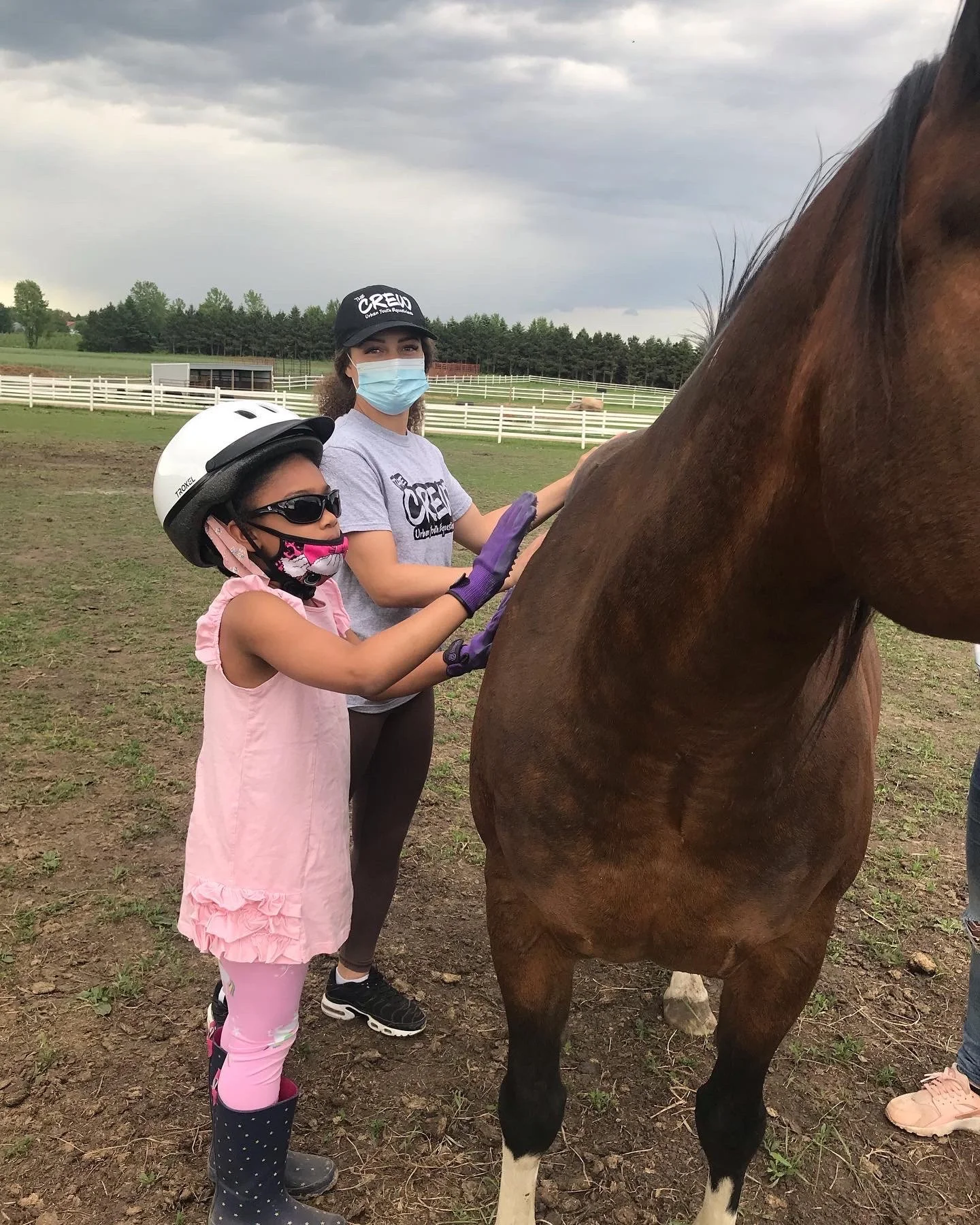 Child wearing a helmet and sunglasses with an adult in a mask grooming a brown horse outdoors on a farm.
