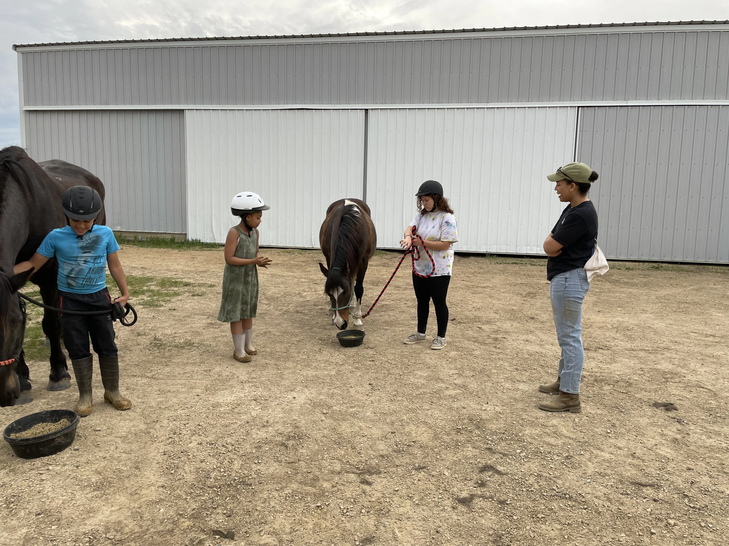 Children and an adult with horses at a rural setting, wearing helmets and casual attire, standing on dirt ground beside a stable building.