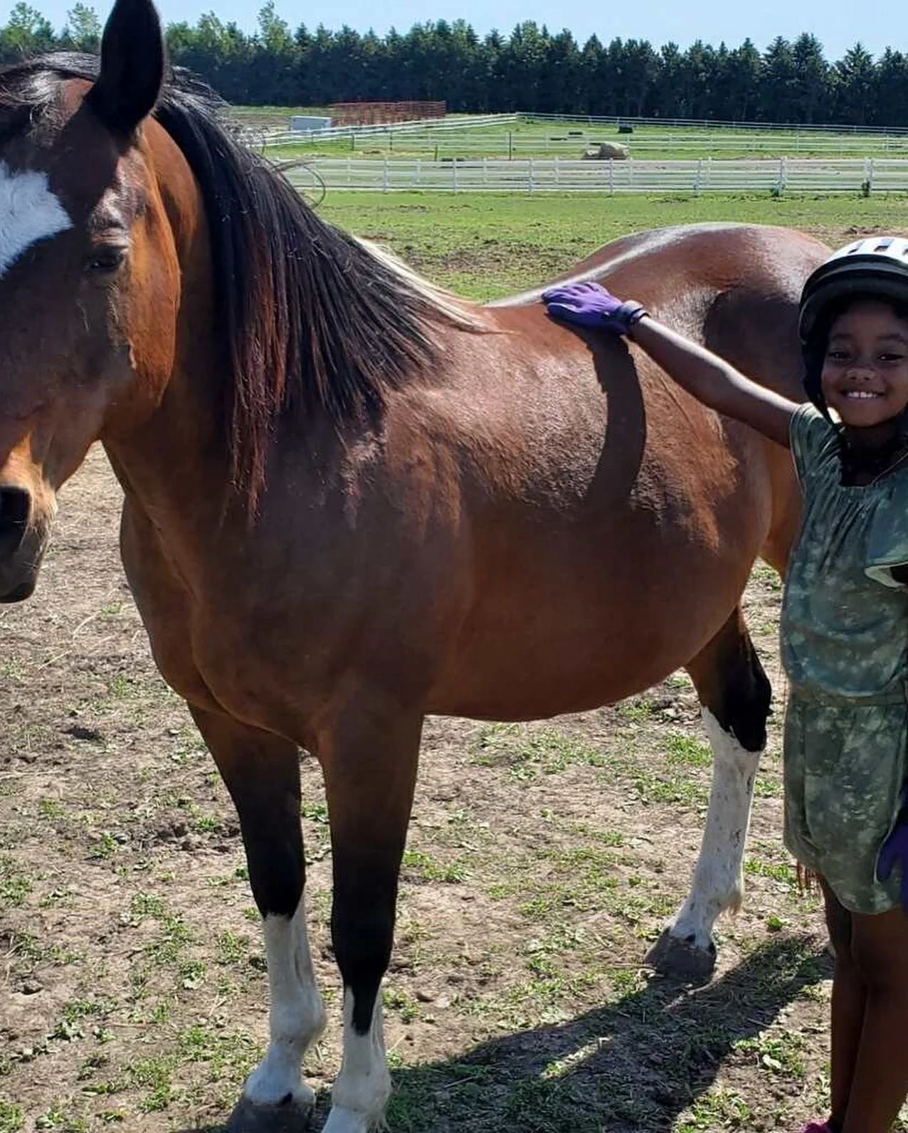 A person in a helmet and gloves petting a brown horse in an outdoor setting.